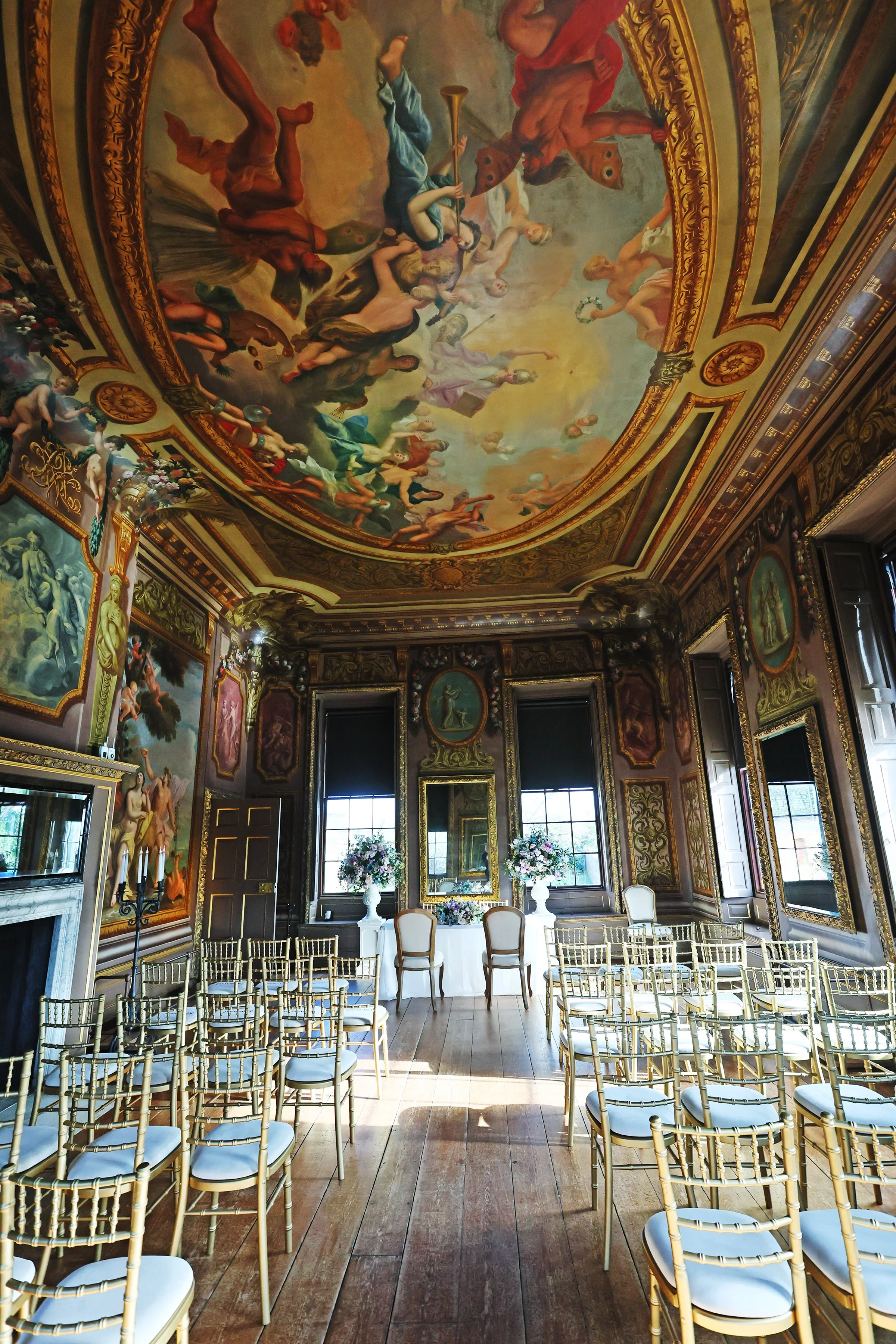 A decorated indoor space with ornate gold wall moldings, paintings, and a vaulted ceiling featuring a large classical painting. The room has wooden floors, tall windows with dark frames, and a table surrounded by gold chairs arranged in rows, likely 