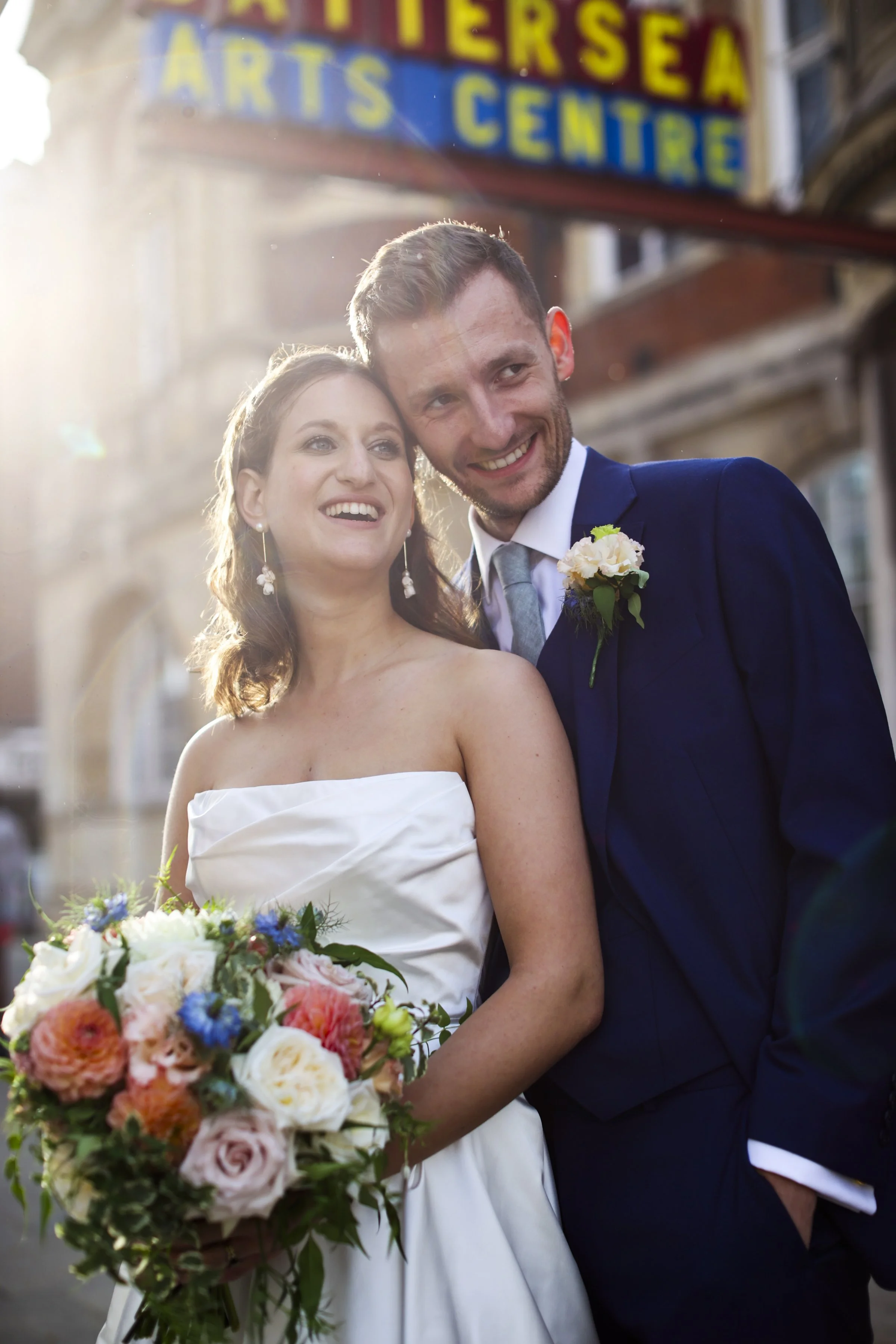 A smiling bride and groom on their wedding day, with the bride holding a large bouquet of colorful flowers, standing outdoors with a sign for the Art Institute of Seattle Arts Center in the background.