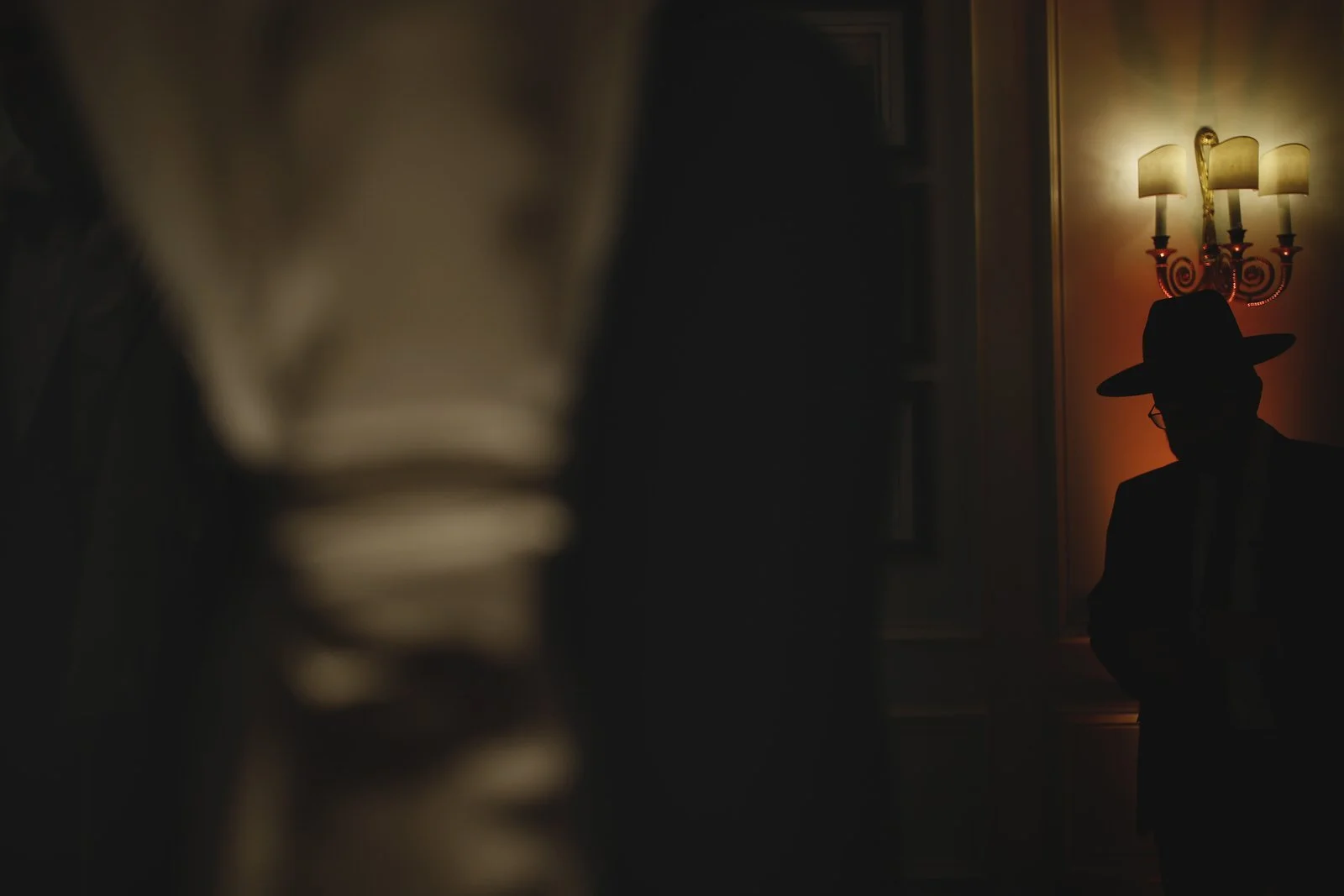 Person wearing a hat and glasses standing in a dimly lit room with a wall sconce