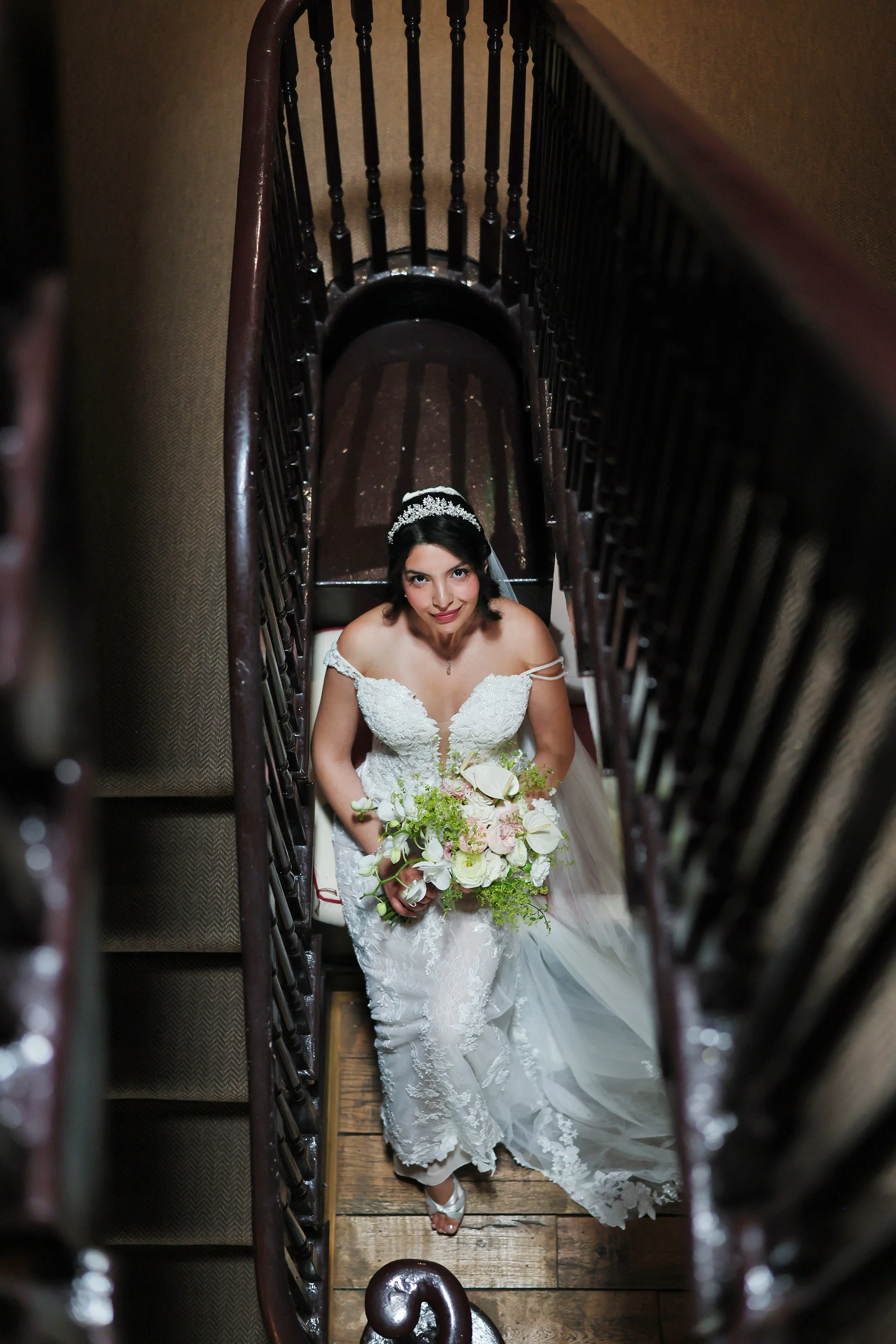 From above view of a bride standing on a staircase, holding a floral bouquet, wearing a white wedding dress and a tiara, looking up at the camera.