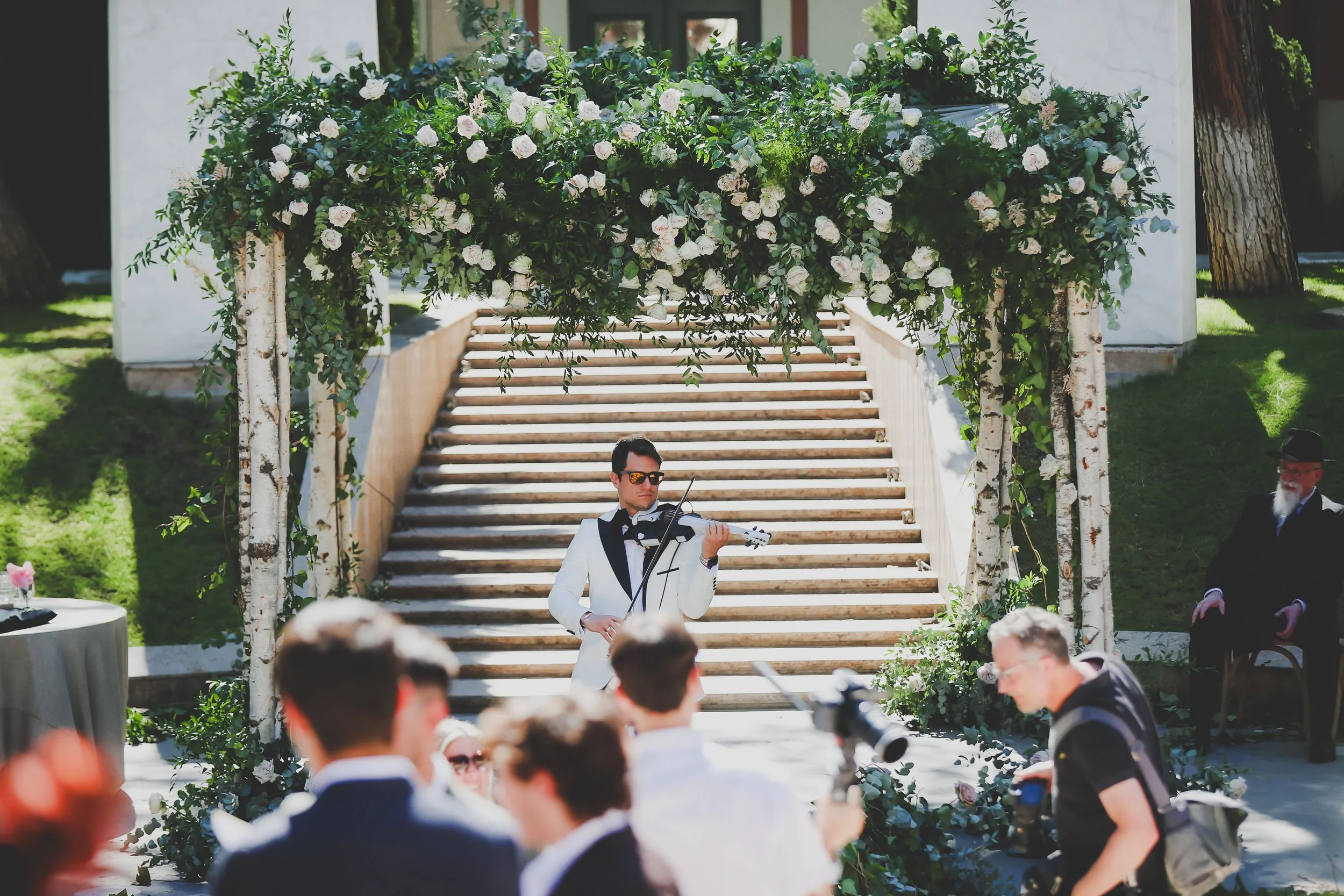 A man in a white tuxedo jacket and black bow tie playing the violin at a wedding ceremony outdoors, with guests seated and a floral archway above.