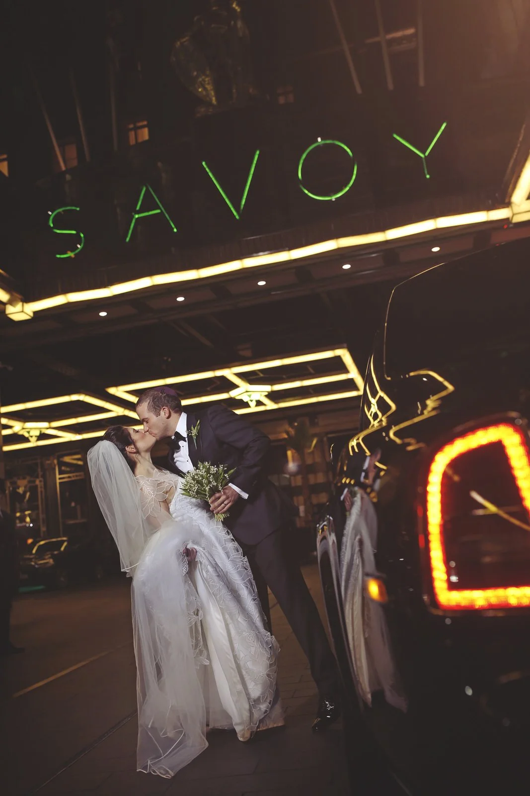 A newlywed couple sharing a kiss in front of a black luxury car at night, with a neon sign above that reads 'SAVOY' and reflects off the car, in an urban setting.