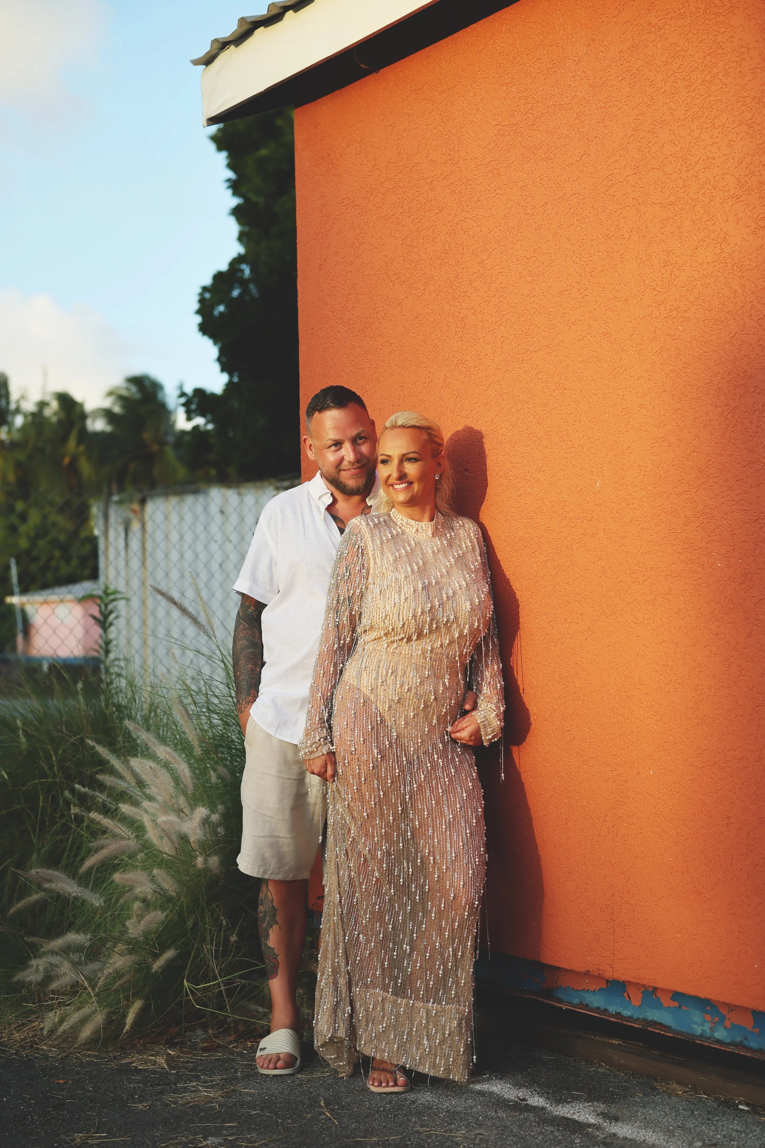 A man and woman stand close together outside, leaning against an orange wall. The man has short dark hair, a beard, and tattoos on his arm, wearing a white shirt and beige shorts. The woman has blonde hair styled back, wearing a shiny, gold, sparkly 