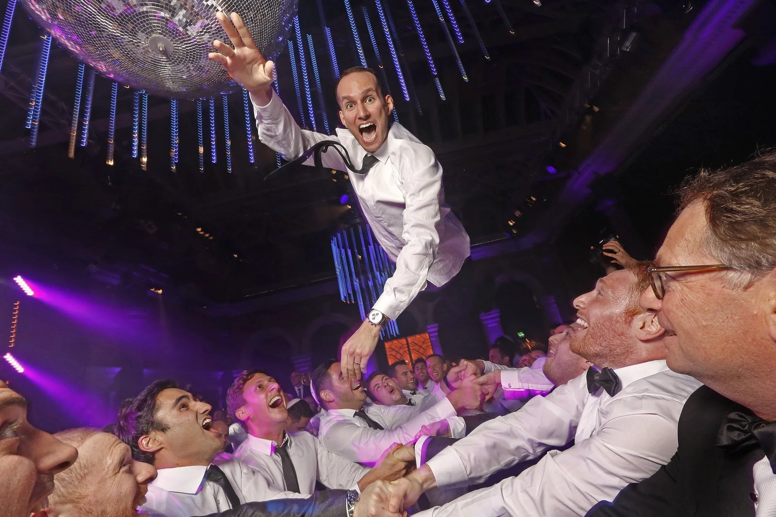 A man in a white shirt and black tie is being lifted by people at a party or wedding reception in a decorated venue with purple and blue lighting, and a large disco ball overhead.