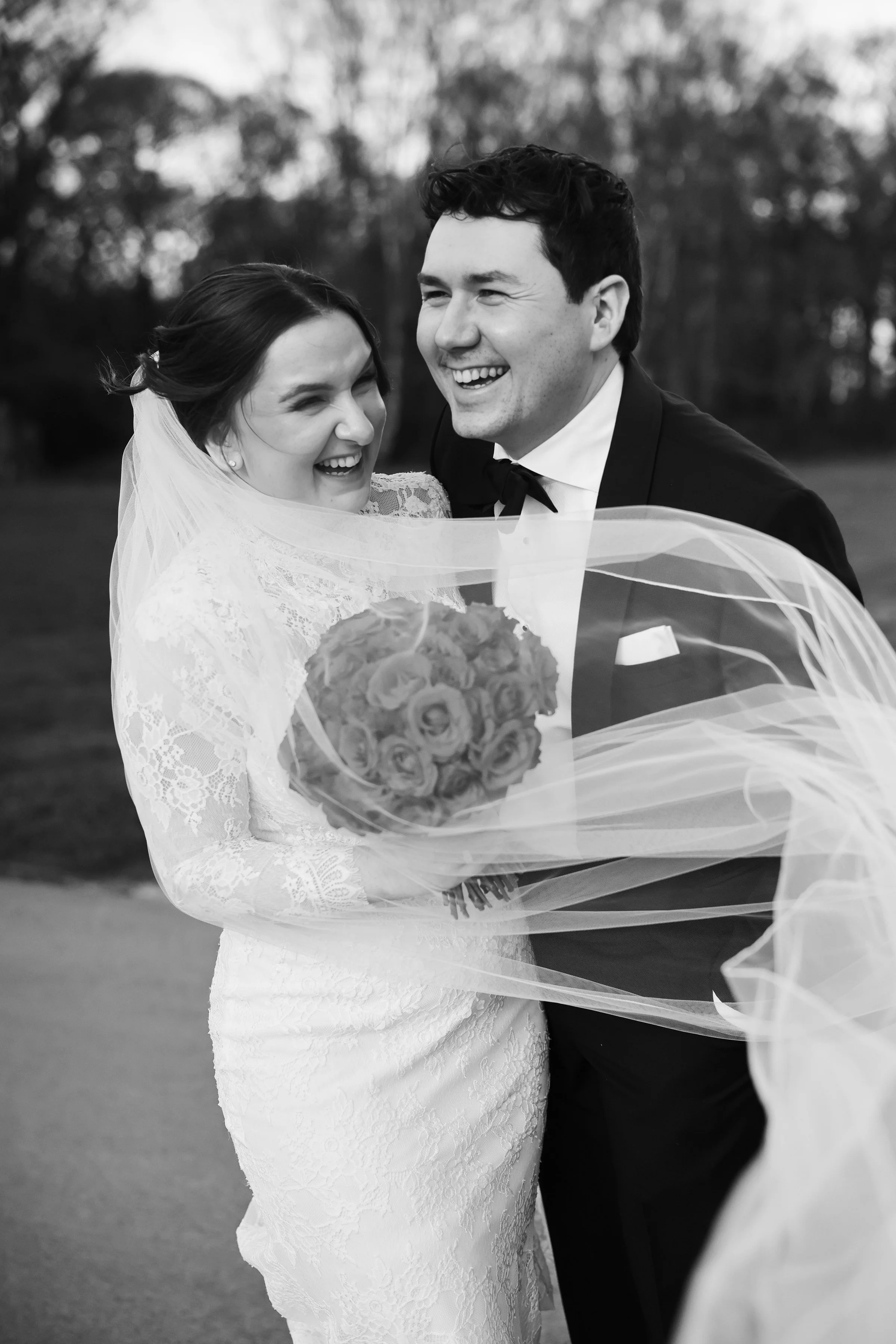 A black-and-white photo of a bride and groom smiling and laughing together, holding a bouquet of flowers wrapped in tulle, outdoors with trees in the background.