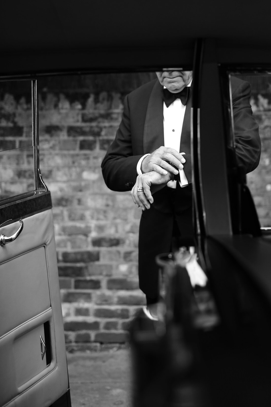 Black and white photograph of a man in a tuxedo, captured from a low angle reflected in a mirror, with a brick wall background.