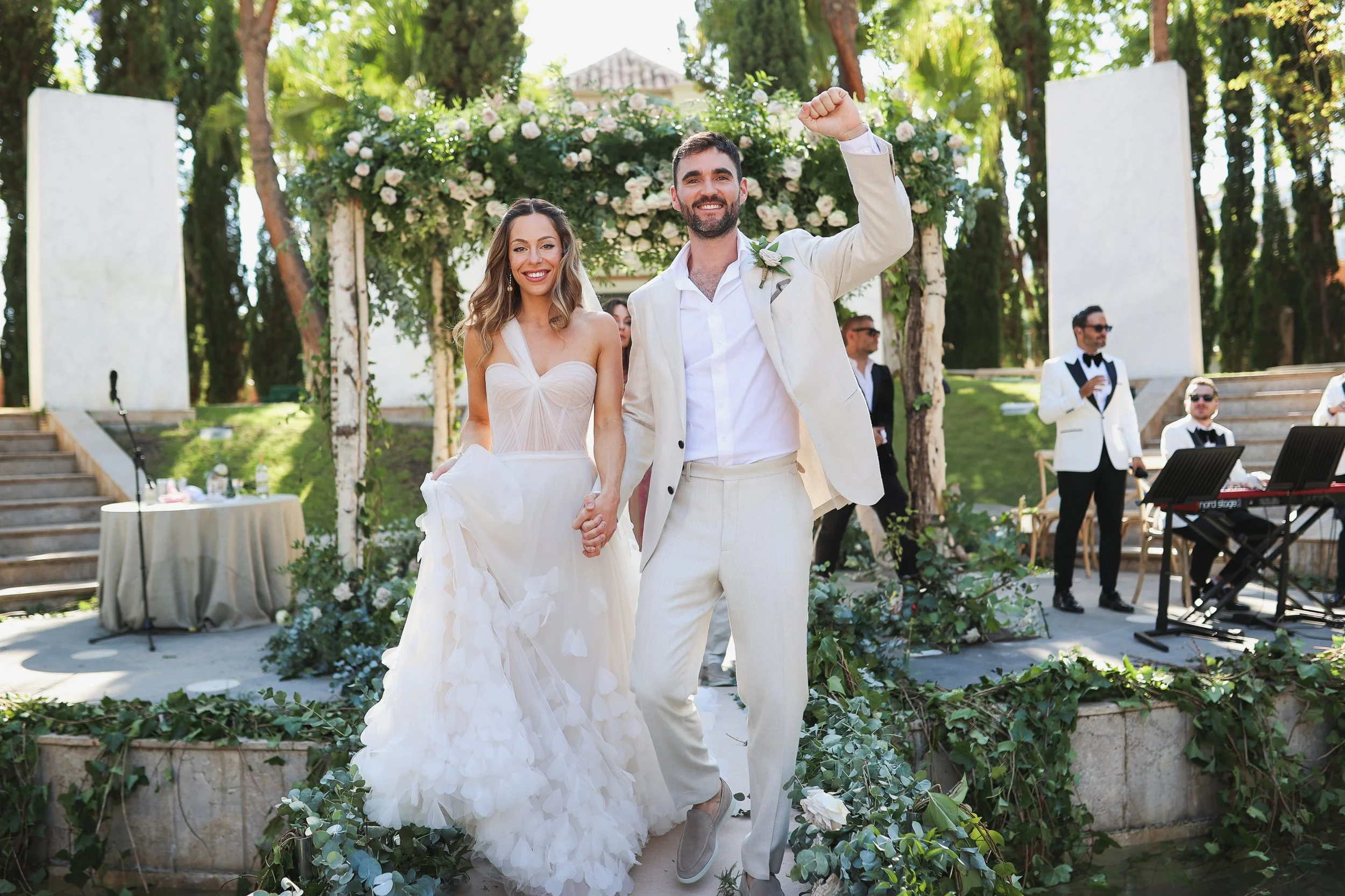 A newlywed couple holding hands and celebrating at outdoor wedding ceremony with floral arch, surrounded by trees and guests, during daytime.