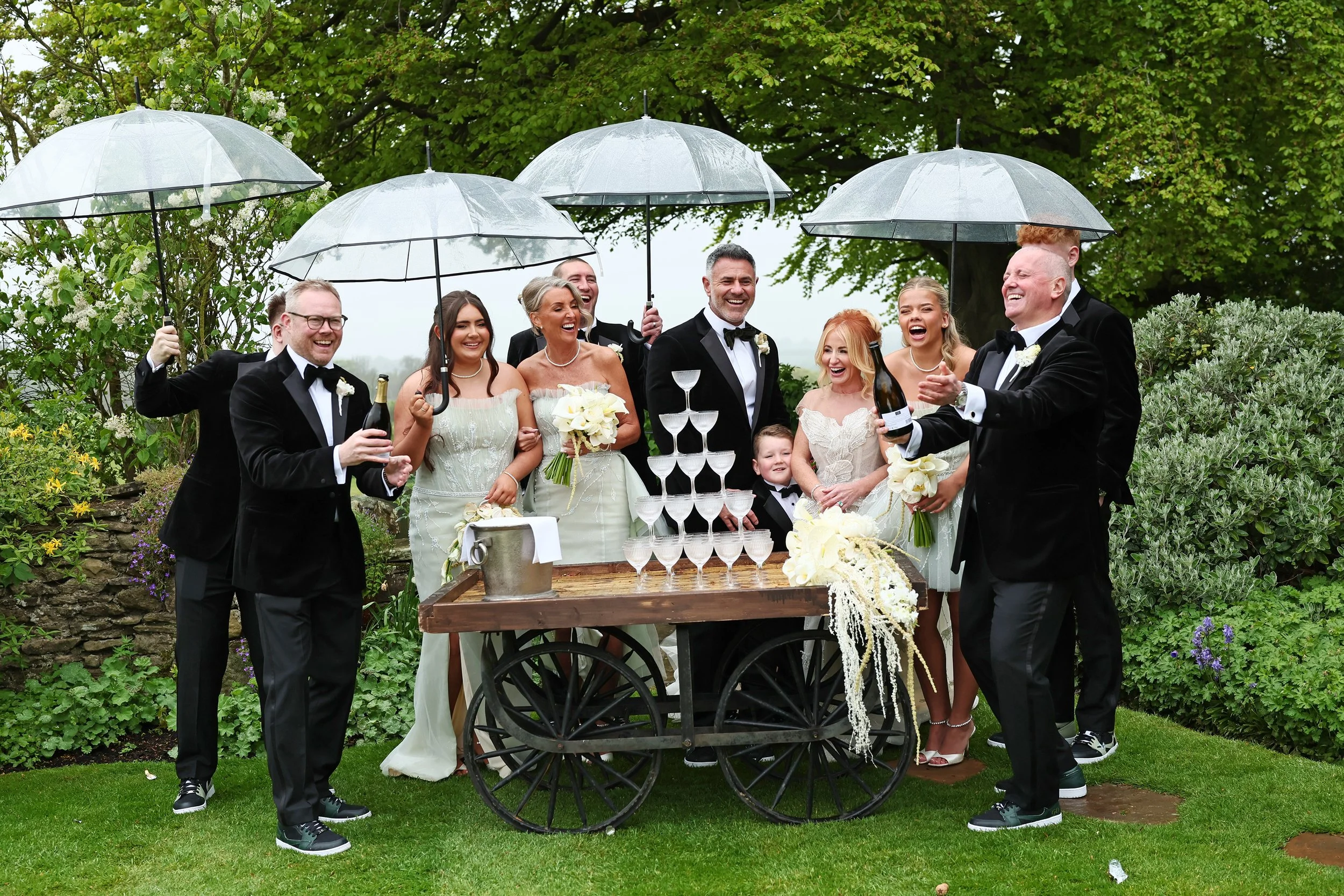 A group of people in wedding attire celebrating outdoors under transparent umbrellas. They are smiling and standing behind a table with a champagne tower. The scene is set in a lush garden with green trees and flowering plants.