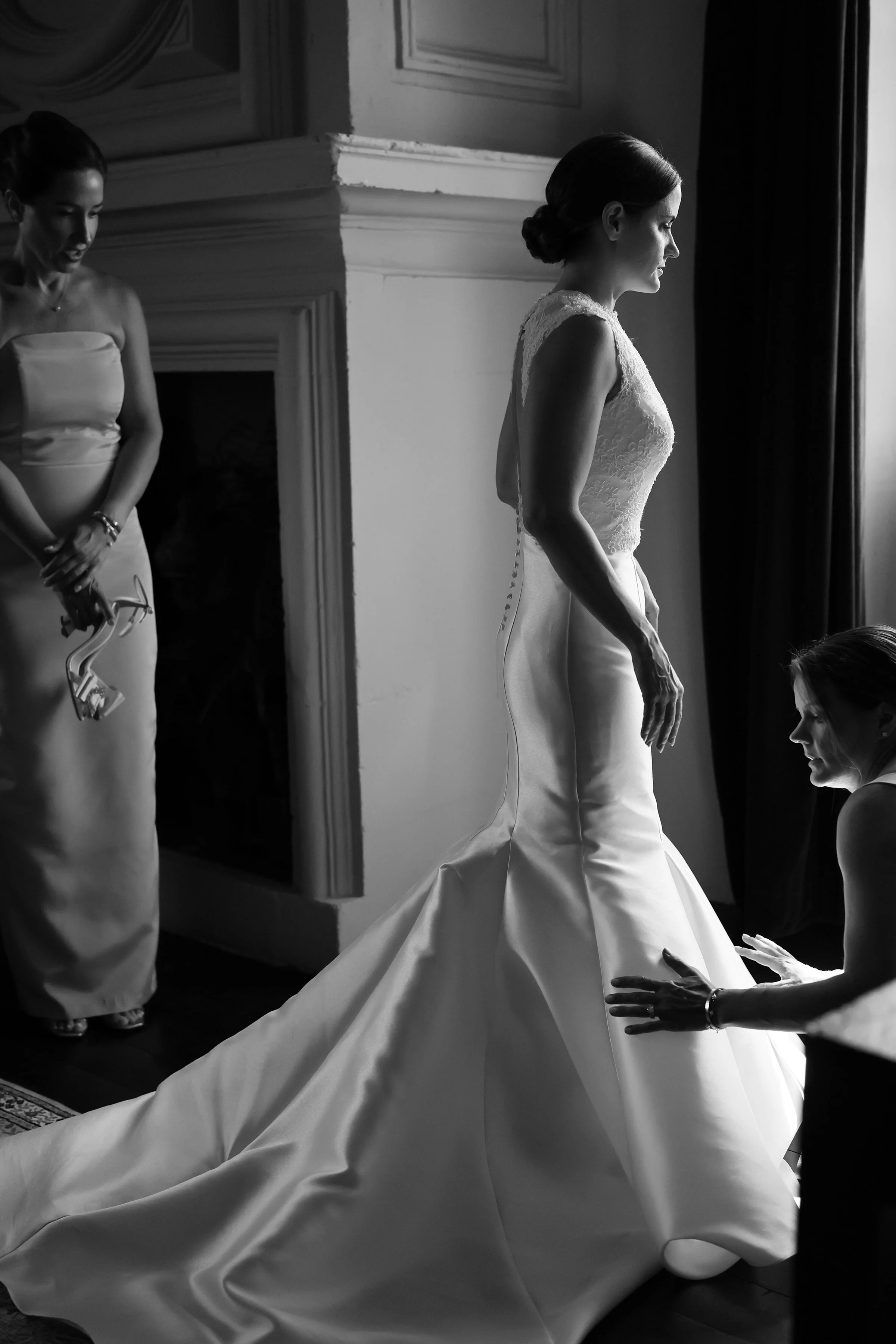 Two women, one in a white wedding dress and the other in a white strapless gown, preparing for a wedding by adjusting the dress in a room with a large window and ornate molding.