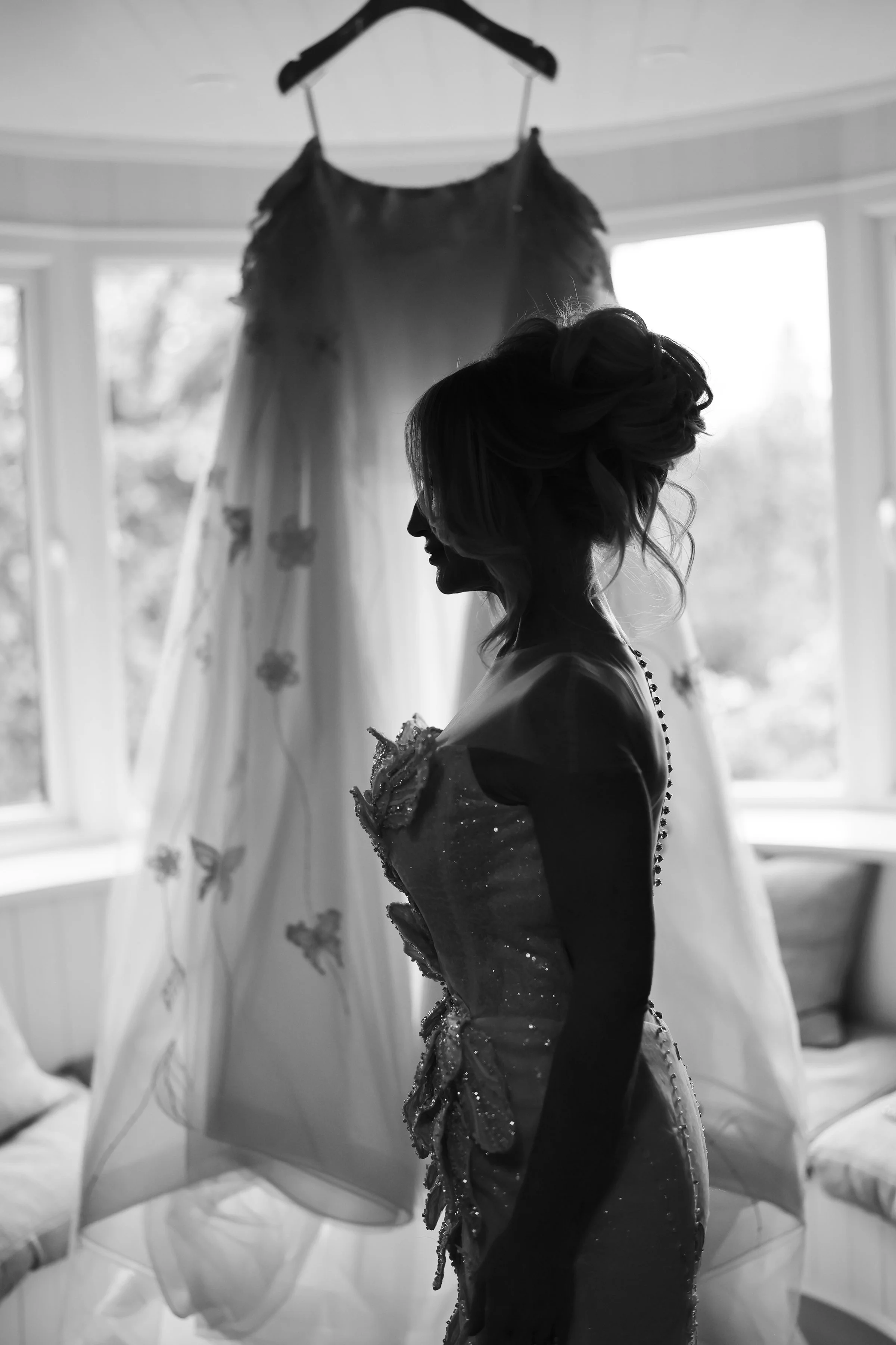 A woman with styled hair wearing a dress with intricate details, standing in profile in front of a window with a wedding dress hanging behind her.