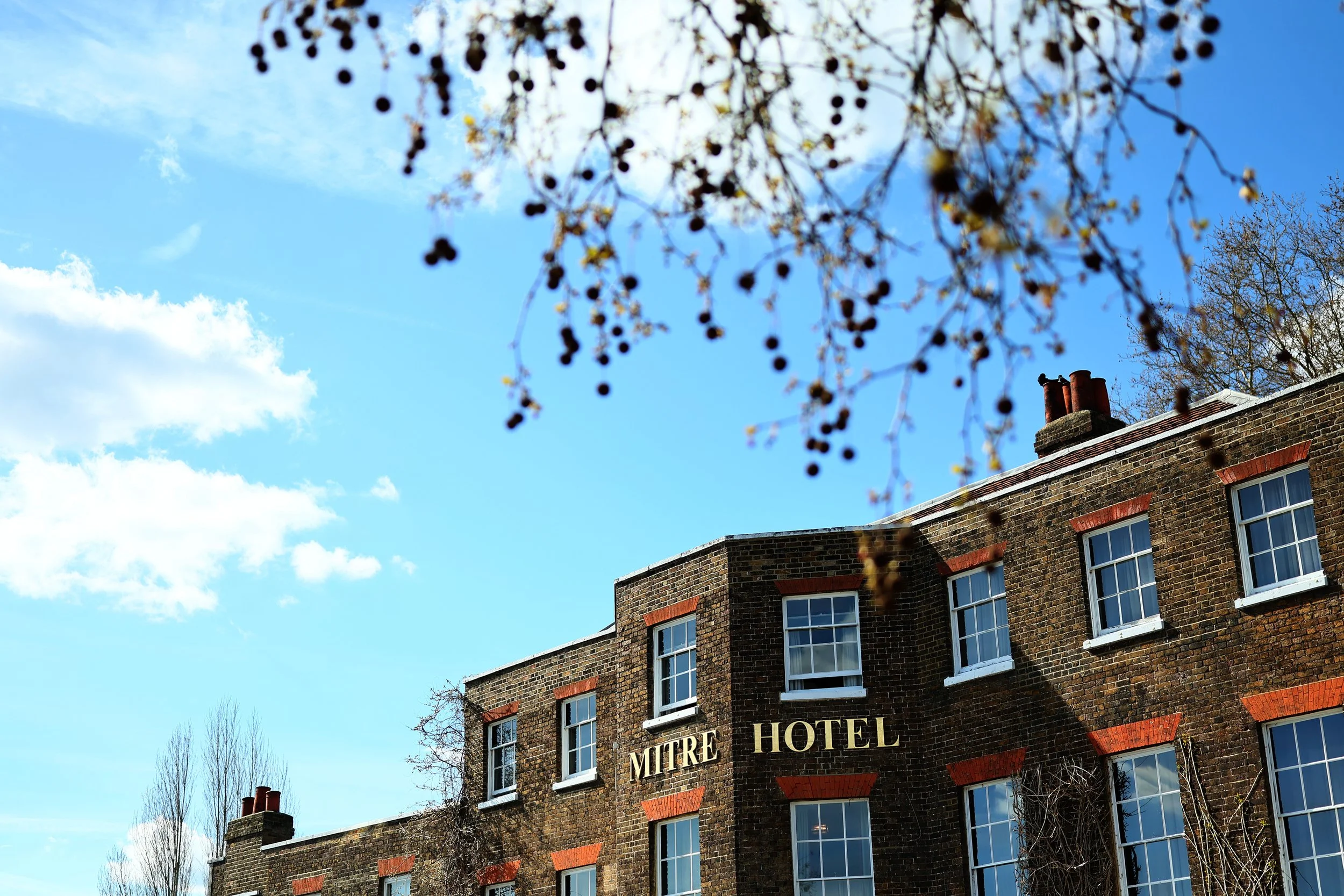 A multi-story brick building with a sign that reads "MITRE HOTEL" on the facade. The top of the building has several chimneys, and there are several windows with white frames. The sky is blue with some clouds, and the photo includes tree branches in 