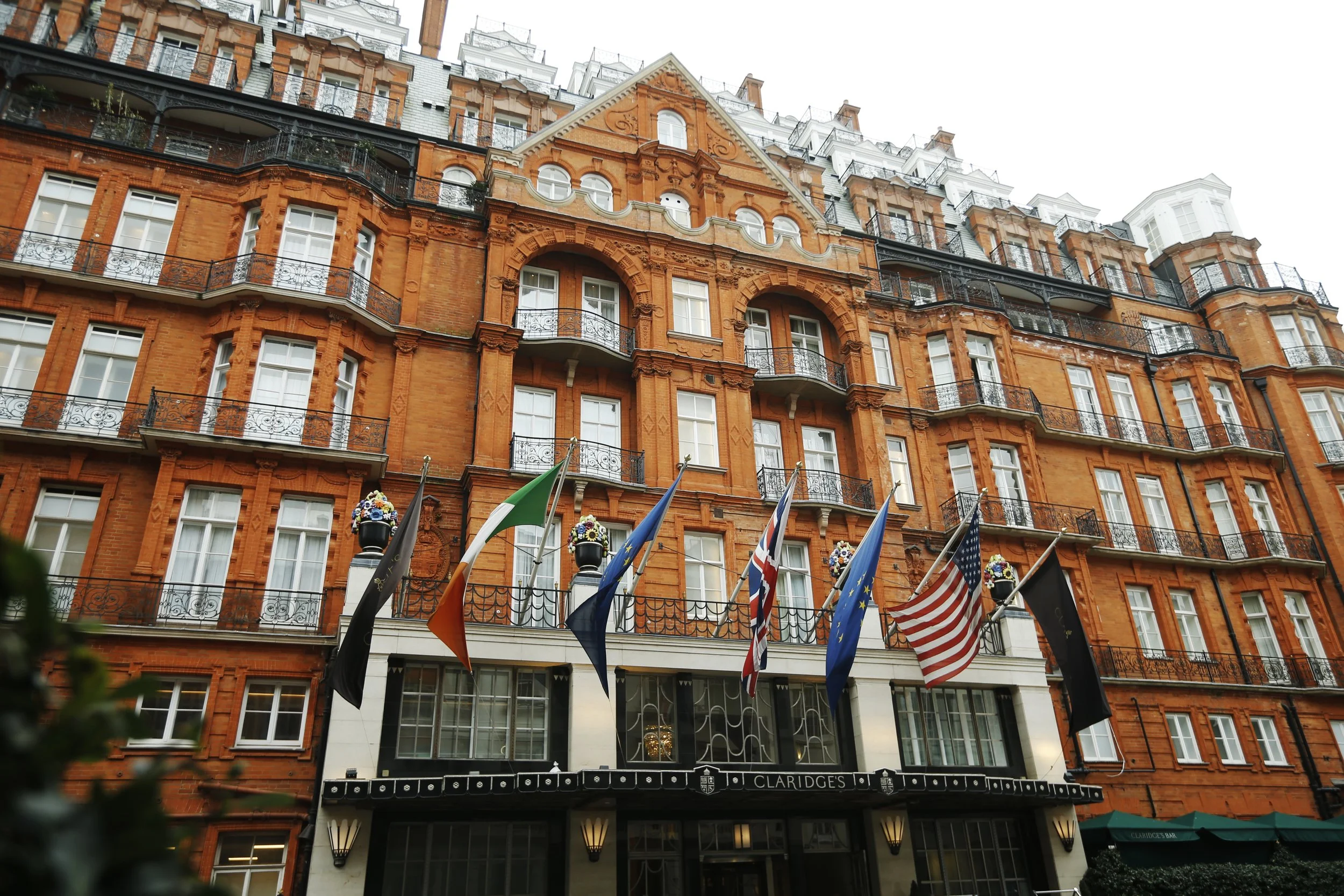 A red brick building with multiple floors, Victorian architecture, and black wrought iron balconies. Flags are hanging outside, and there is a sign that reads 'Claridge's' at the entrance.