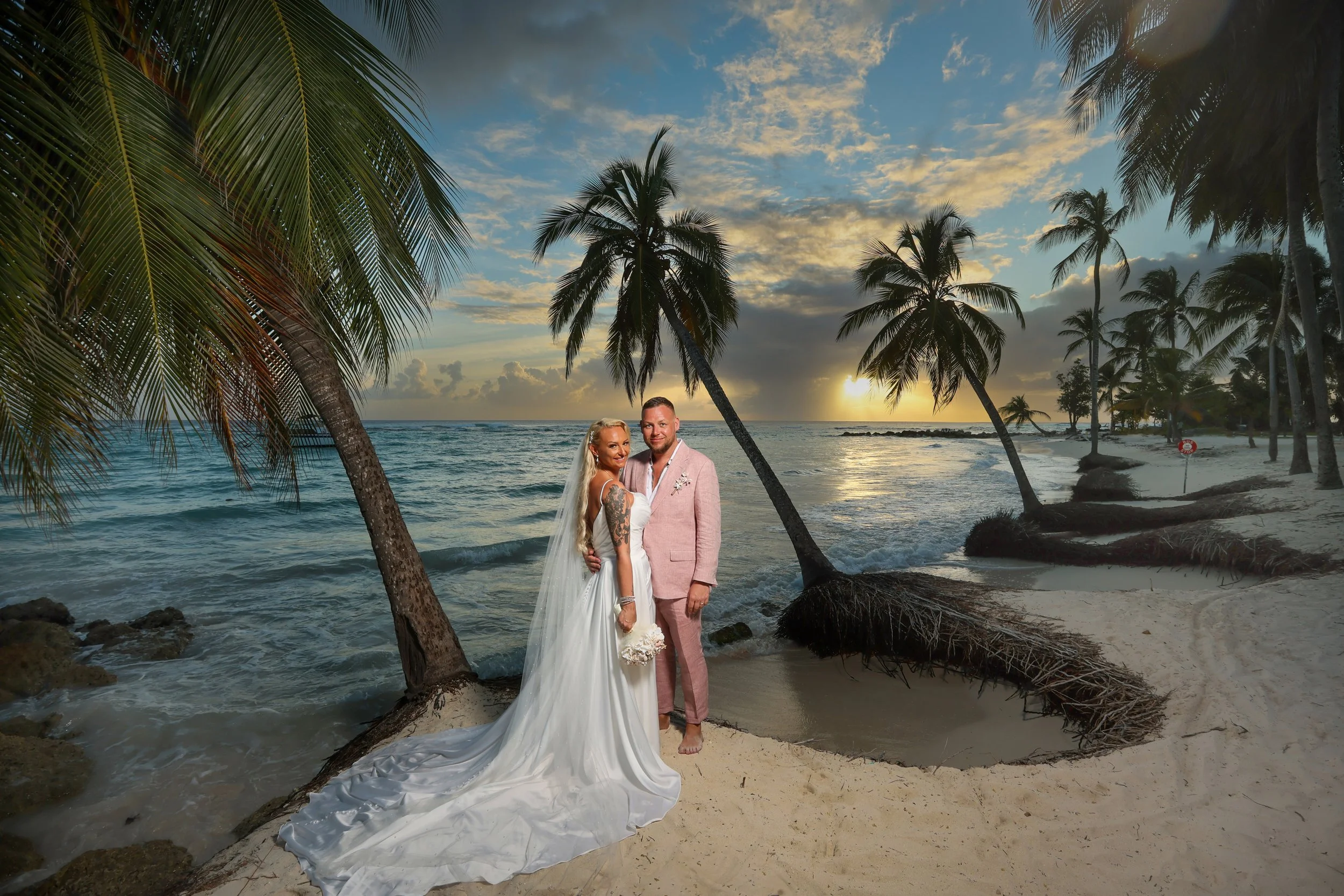 A couple dressed in wedding attire standing on a sandy beach at sunset, surrounded by tall palm trees over calm ocean waters.