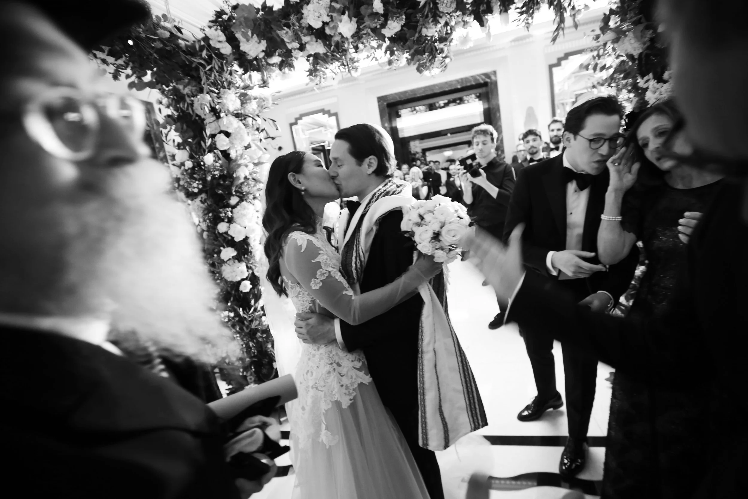 A black and white photo of a wedding ceremony where a bride and groom are sharing a kiss under a floral arch, surrounded by guests in formal attire.