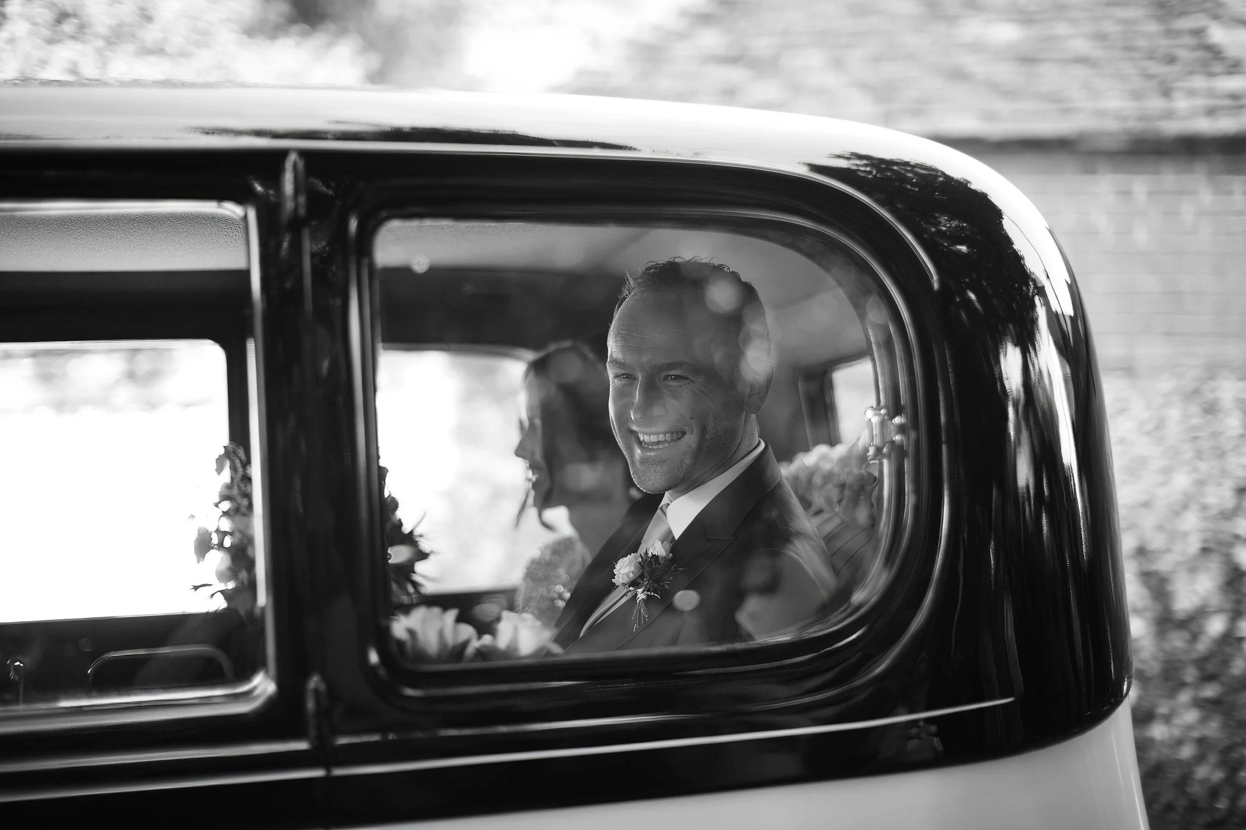 A smiling man in a suit with a boutonniere sitting inside a vintage car, with a woman in the background, during daytime.