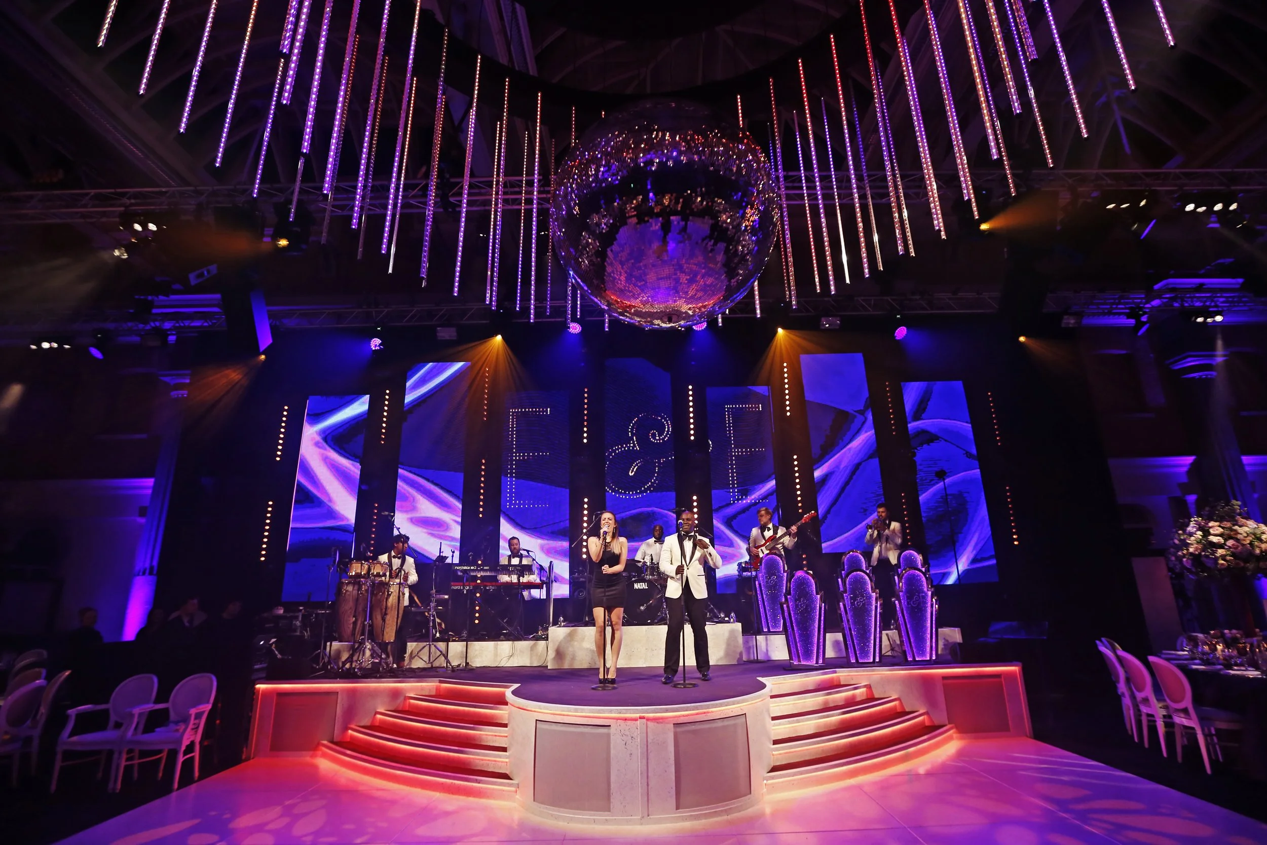 Live band performing on a stage with vibrant purple and pink lighting, crystal ball chandelier overhead, and a large digital screen displaying abstract visuals in the background.