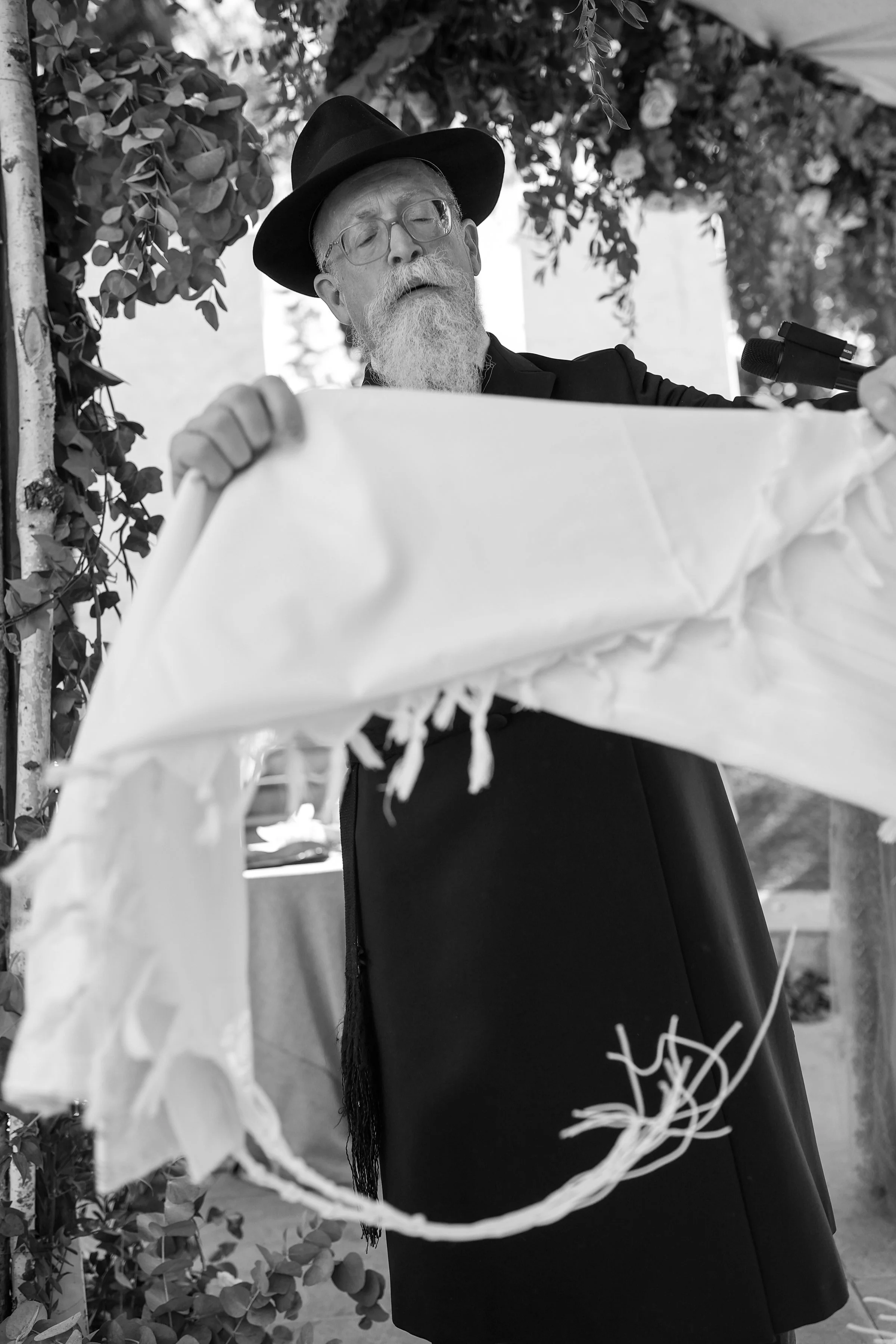 A bearded man with glasses wearing a dark wide-brimmed hat and a dark jacket, standing behind a decorative wreath or hanging ornament made of paper or fabric, with leafy foliage in the background.