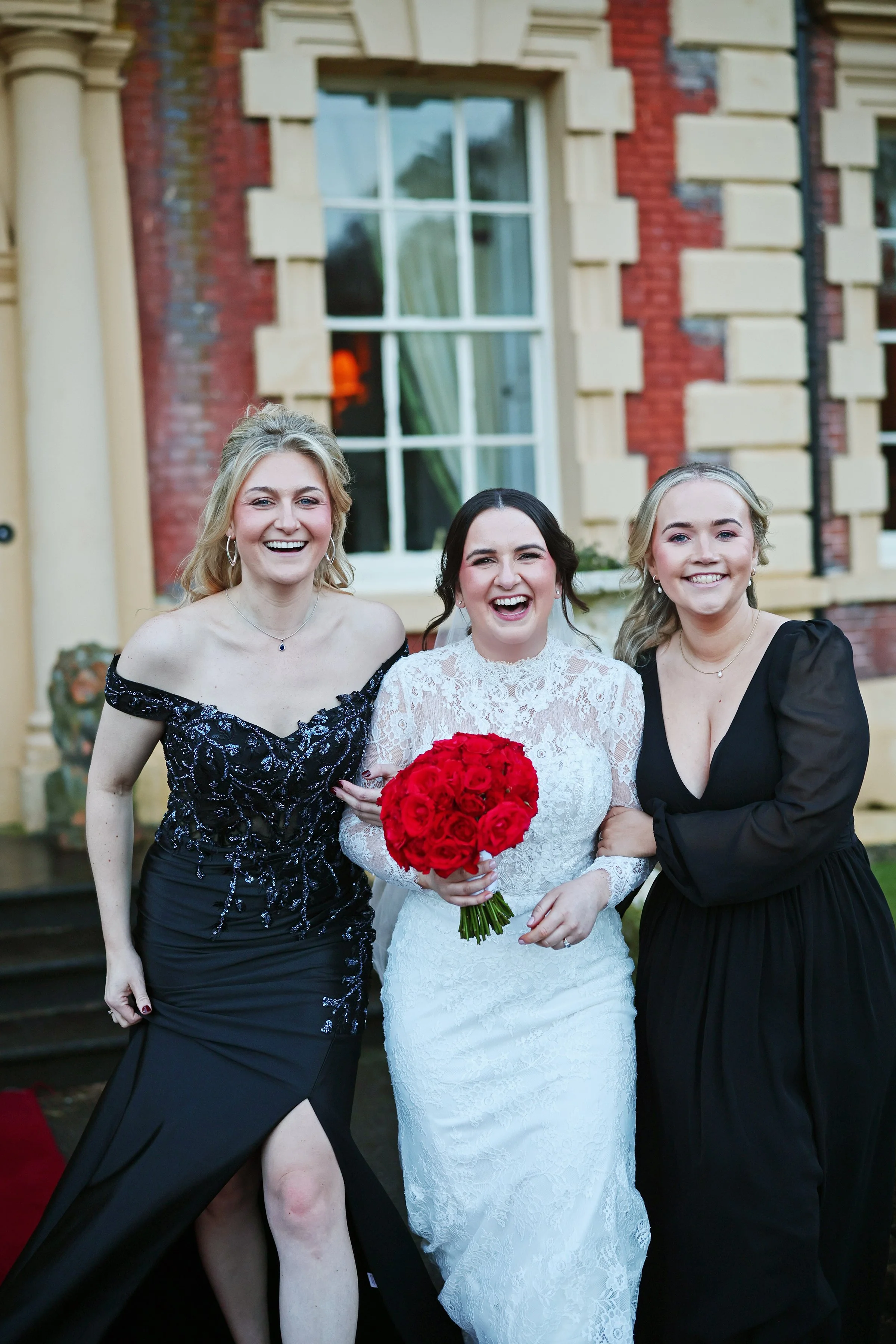 Three women standing together outdoors in front of a brick building, smiling for the camera. The woman in the middle is wearing a white lace wedding dress holding a bouquet of red roses. The woman on the left is wearing a black dress with a slit, and