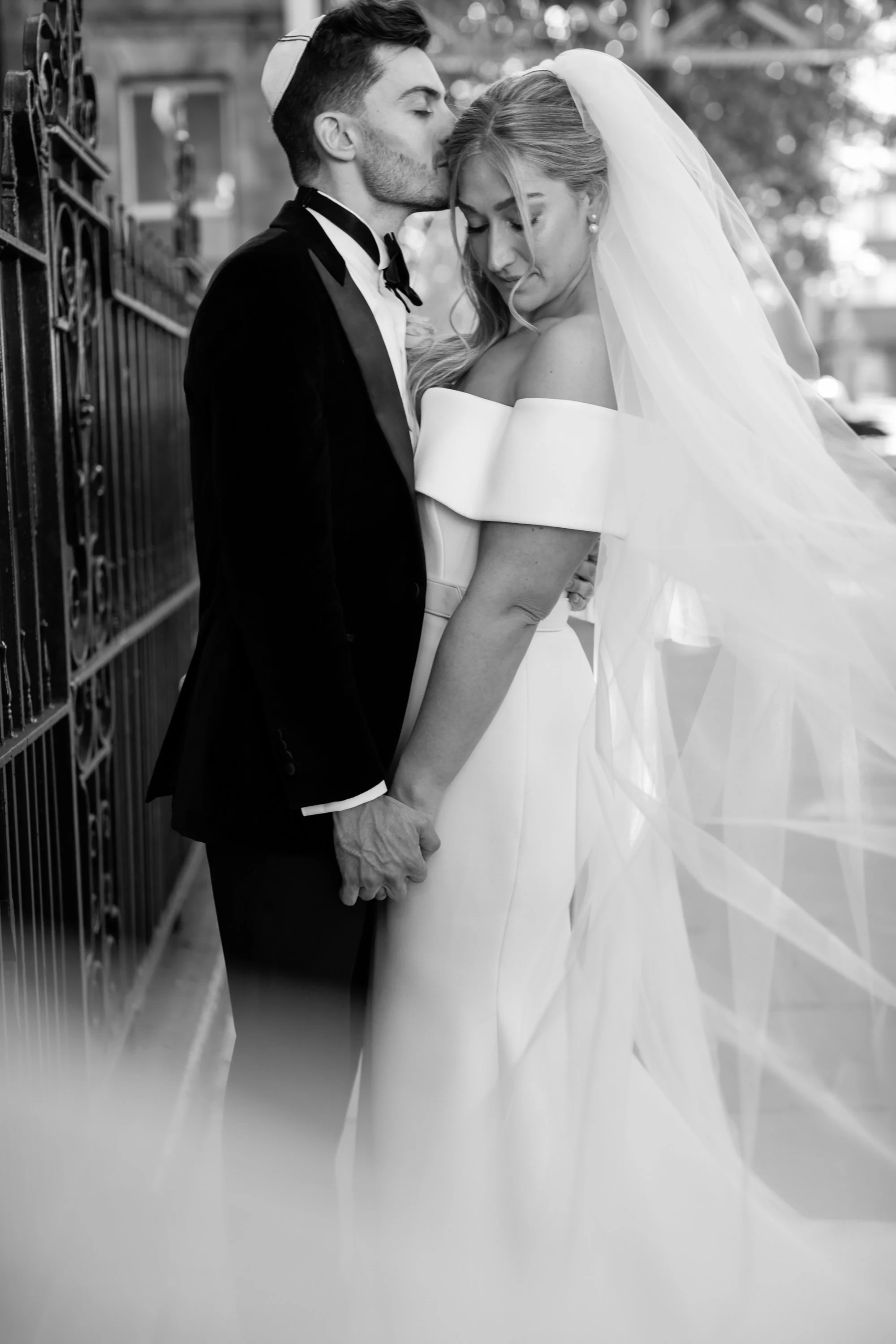 Black and white photograph of a bride and groom in wedding attire, with the groom kissing the bride on the forehead, holding hands, outdoors near a fence with blurred trees in the background.
