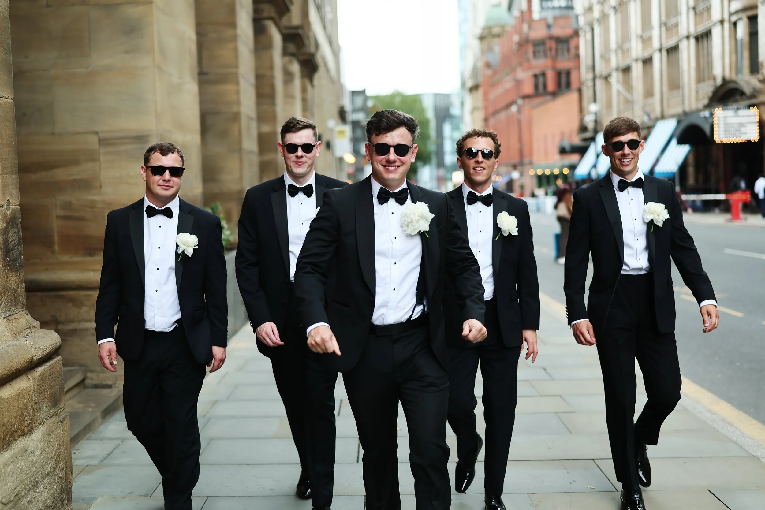 A group of five men dressed in black tuxedos and sunglasses walking on city sidewalk, smiling.