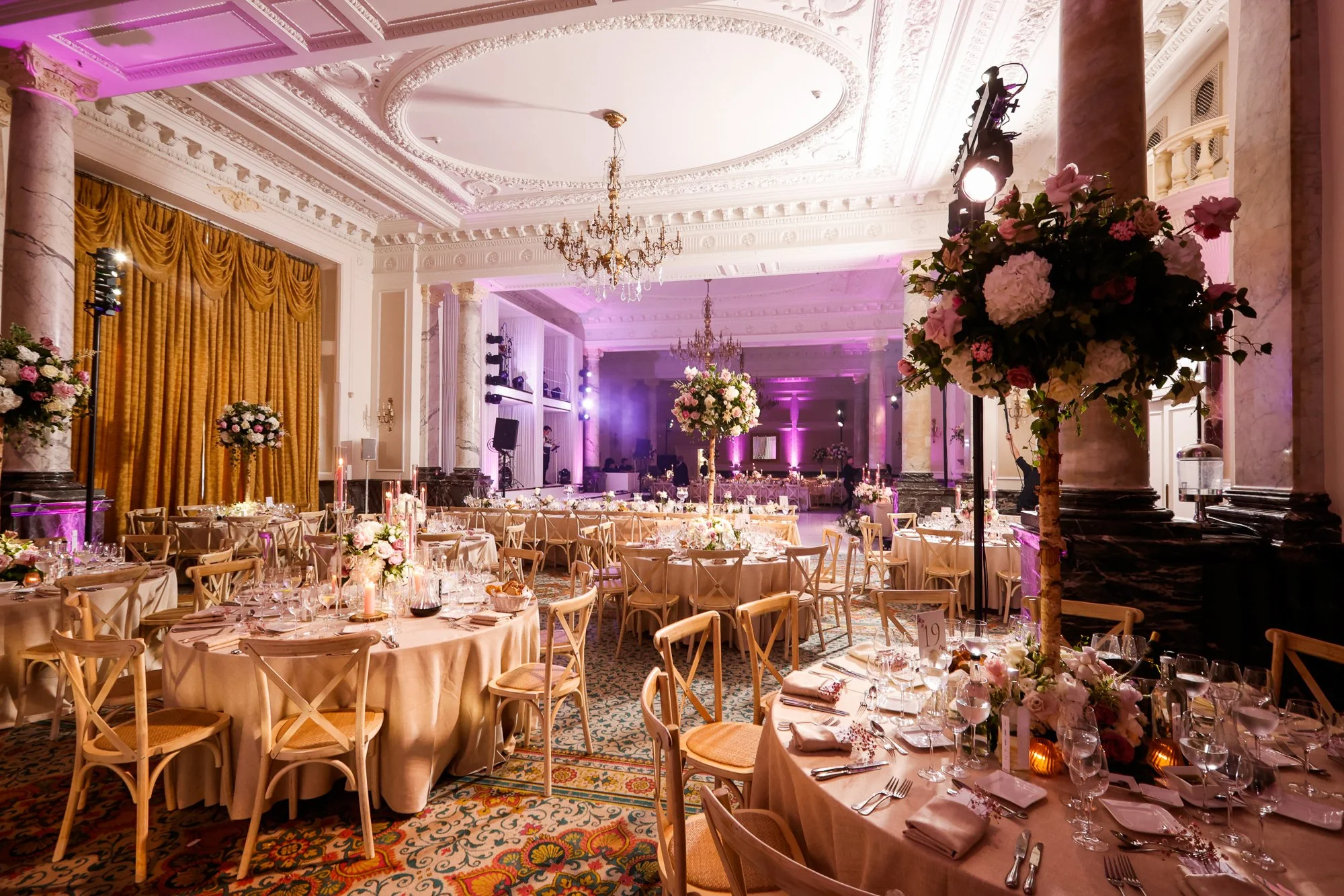 Elegant banquet hall decorated with pink and white flowers, tall floral centerpieces on round tables, gold curtains, marble columns, chandeliers, and purple lighting.