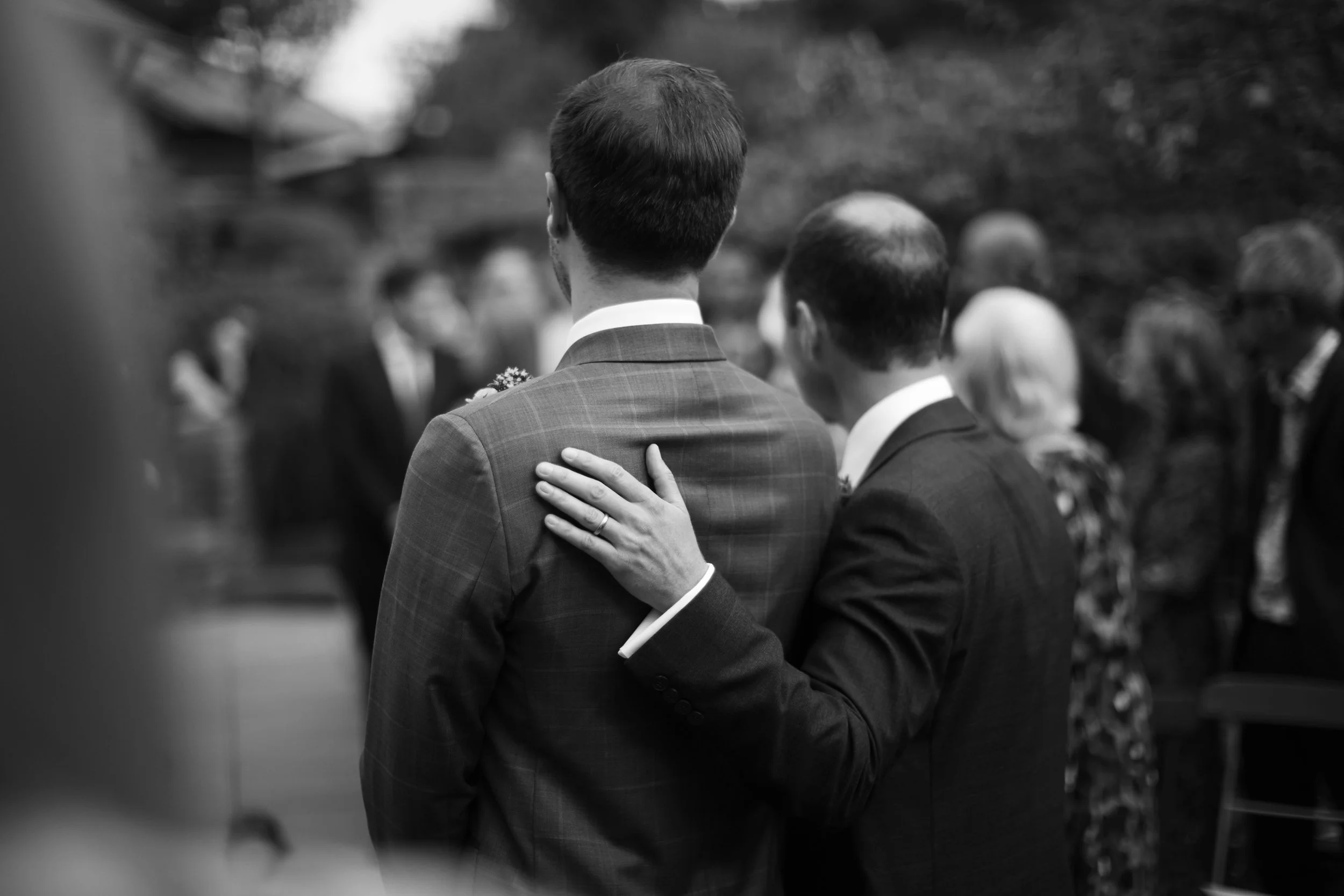 Black and white photo of a groom and a guest embracing at a wedding, with blurred wedding guests in the background.