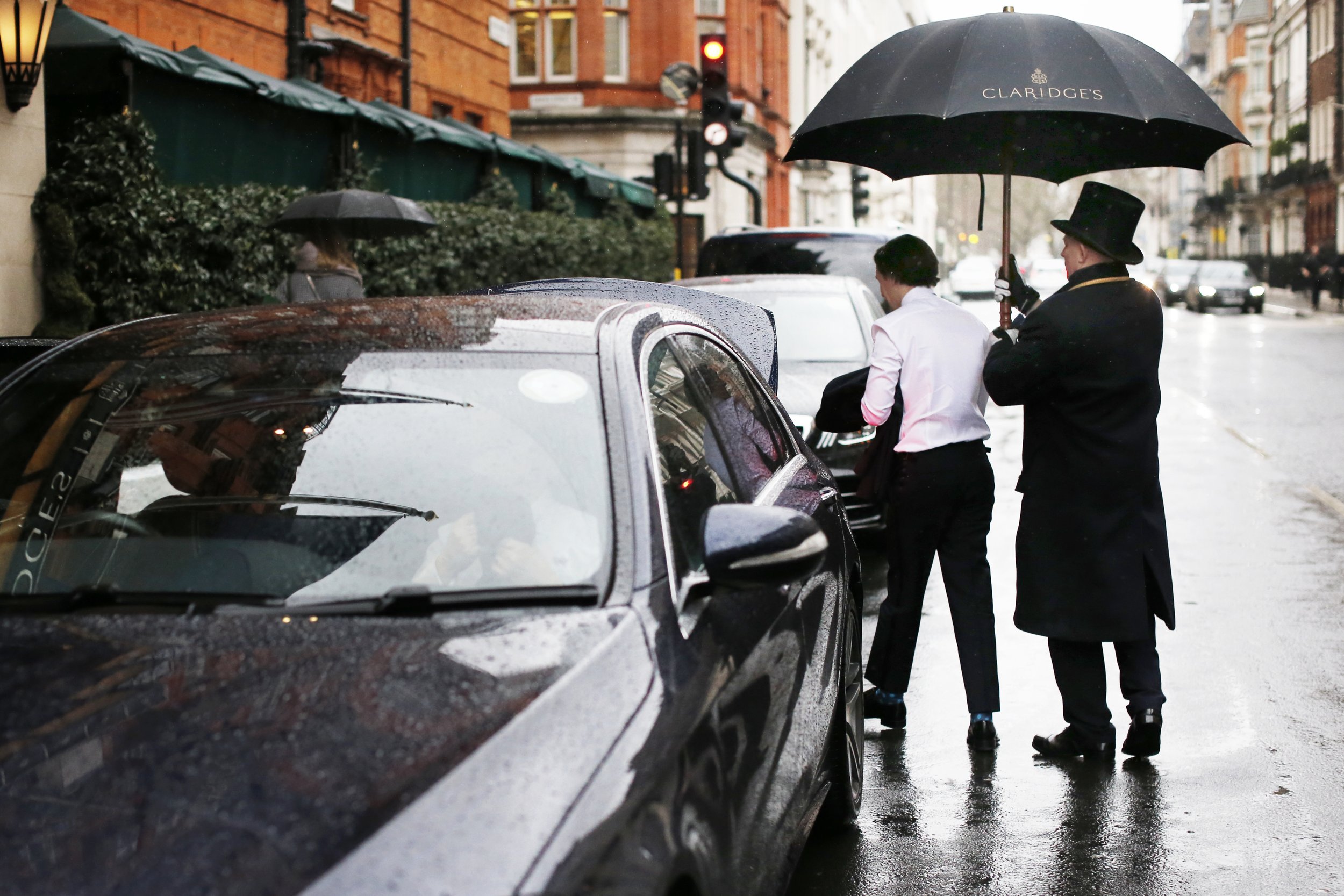 A man with a top hat and black coat holding an umbrella labeled 'Claridge's' walking beside another person on a rainy city street, near parked cars with raindrops on their surfaces.