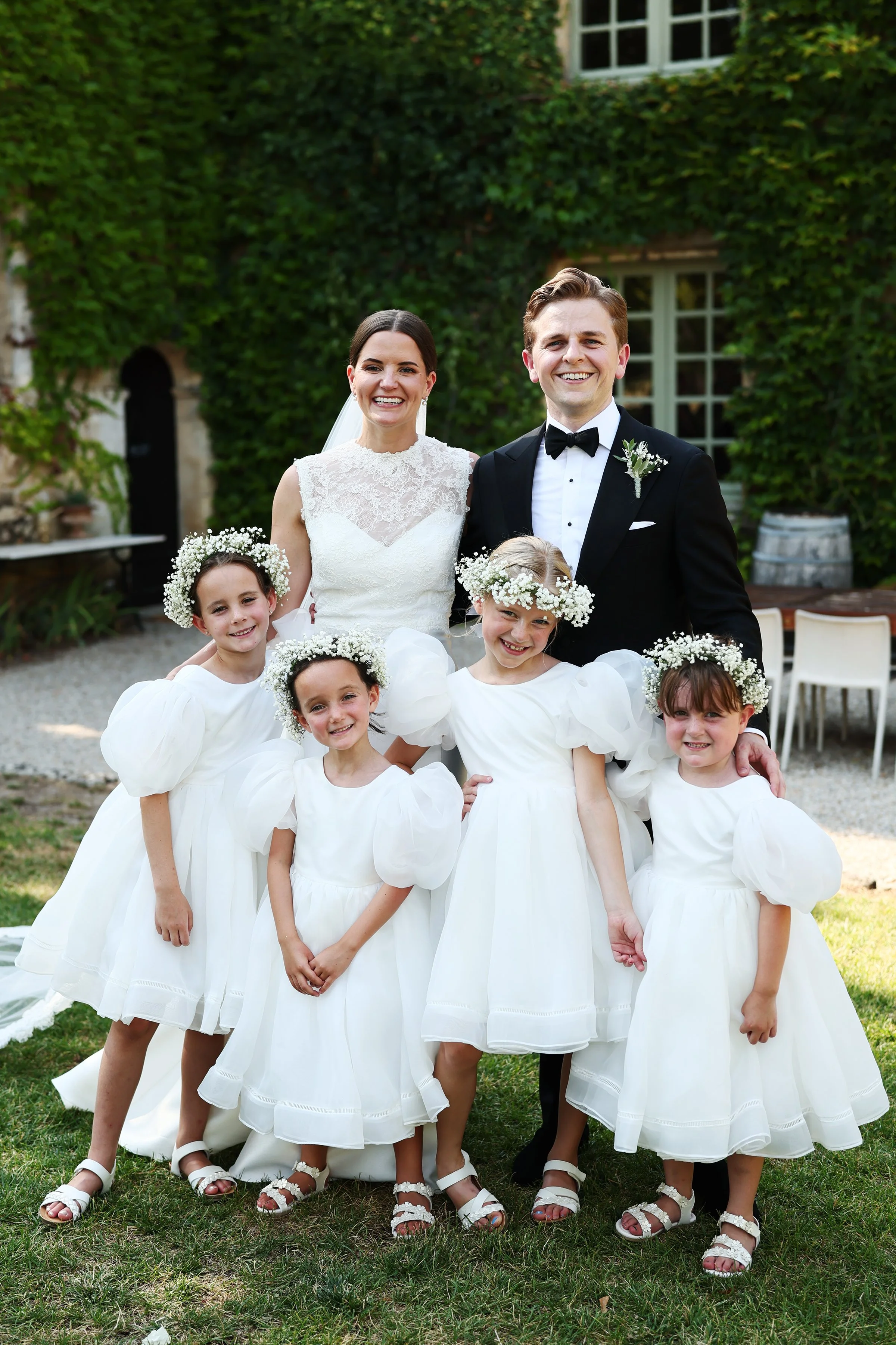 A group of children and a bride and groom smiling outdoors during a wedding celebration. The children are dressed in white dresses with floral headbands, and the bride and groom are in wedding attire.