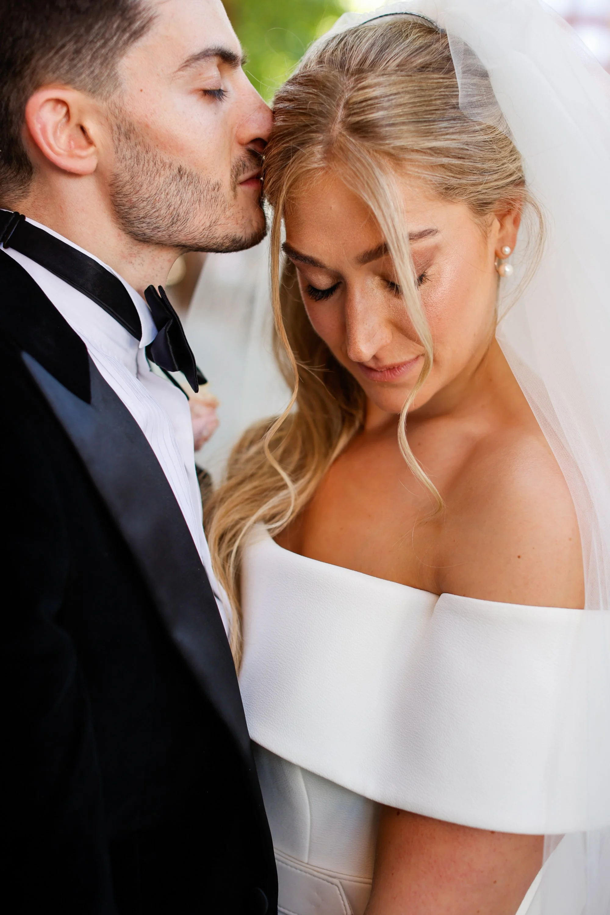 A man and a woman are in a close, intimate moment, with the man kissing the woman's forehead. The man is dressed in a tuxedo with a bow tie, and the woman is wearing a white off-the-shoulder wedding dress with a veil. Both have their eyes closed.