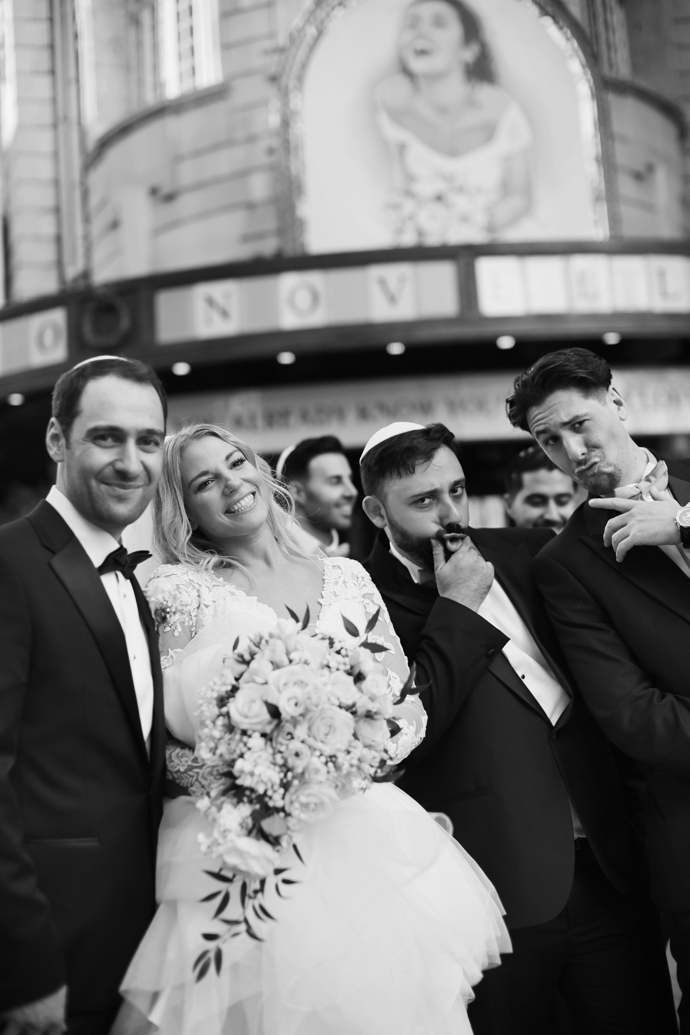 Black and white photo of a wedding party in formal attire, with a church or theater marquee in the background displaying a picture of a woman and the word 