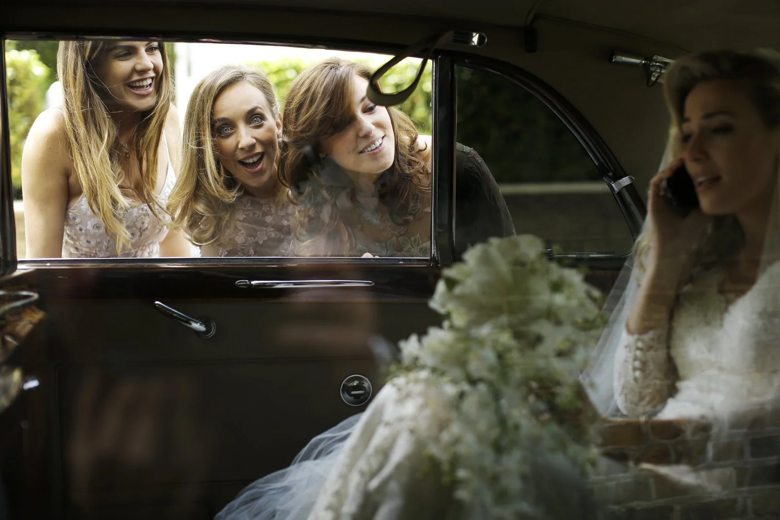 A bride sitting inside a vintage car talking on the phone, with a bouquet of white flowers in the foreground. Outside the car, three women are looking in through the window, smiling and excited.