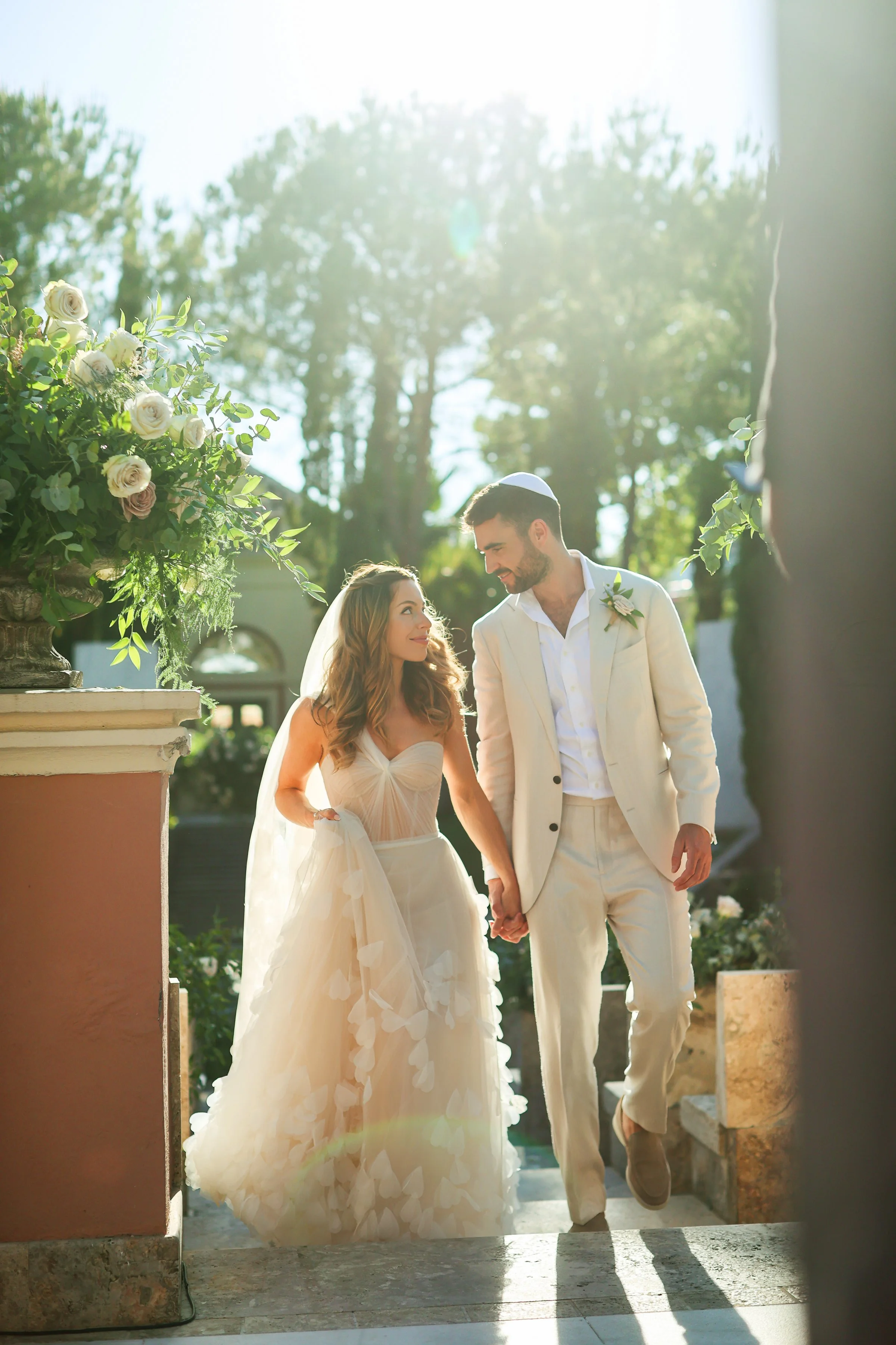 A bride and groom walking hand in hand outdoors during a wedding ceremony, smiling and looking at each other. The bride is wearing a strapless white wedding gown with floral appliques, and the groom is dressed in a white suit with a boutonniere. Ther