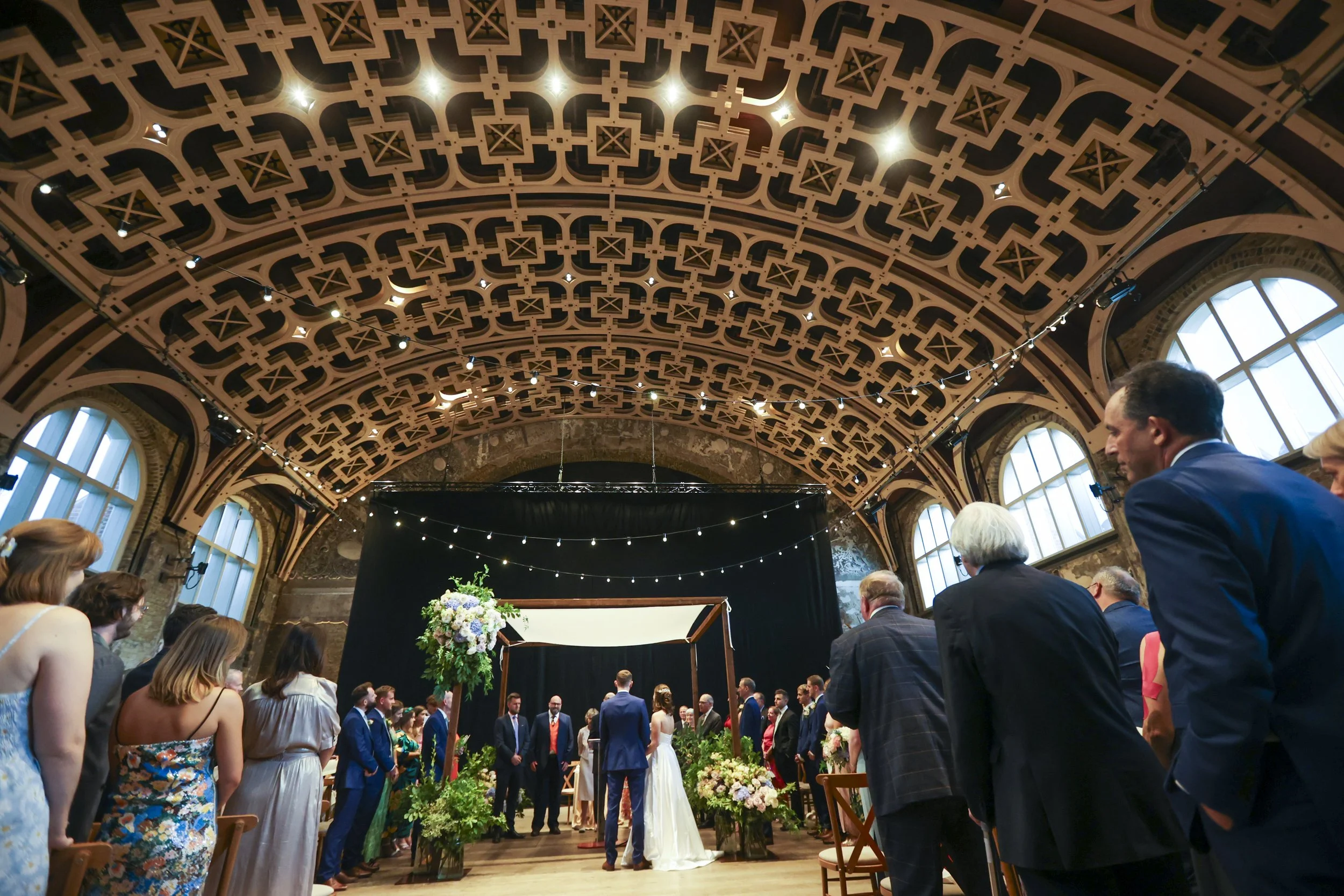 People attending a wedding ceremony inside a grand hall with a decorated ceiling and large windows, standing in a semi-circle around the couple.