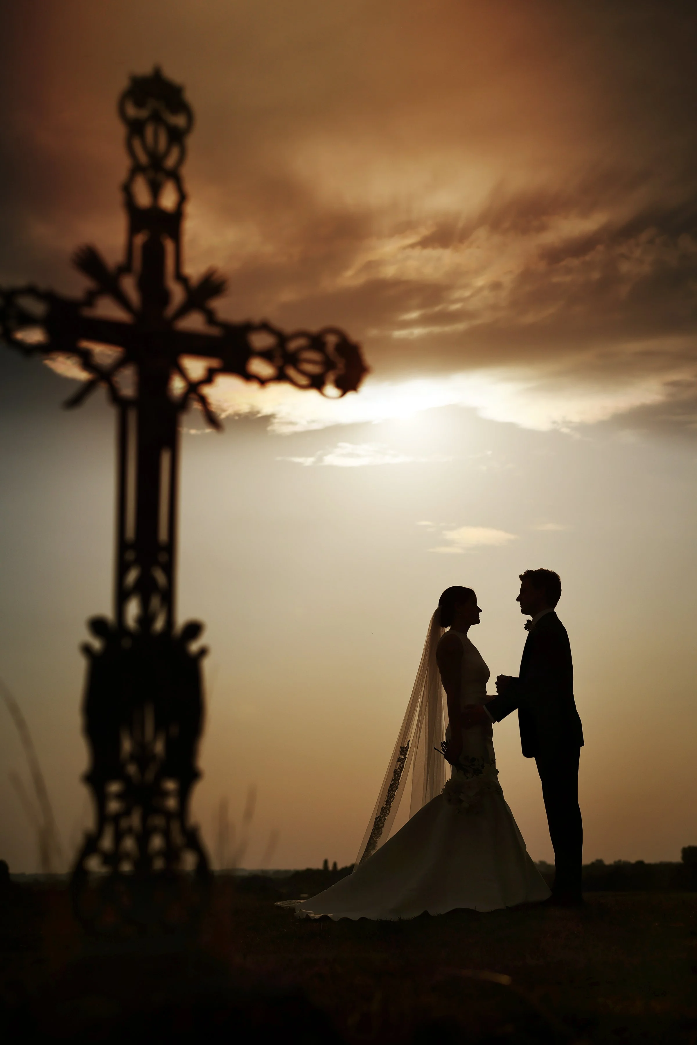 Silhouette of a bride and groom holding hands during sunset with a decorative cross in the foreground.