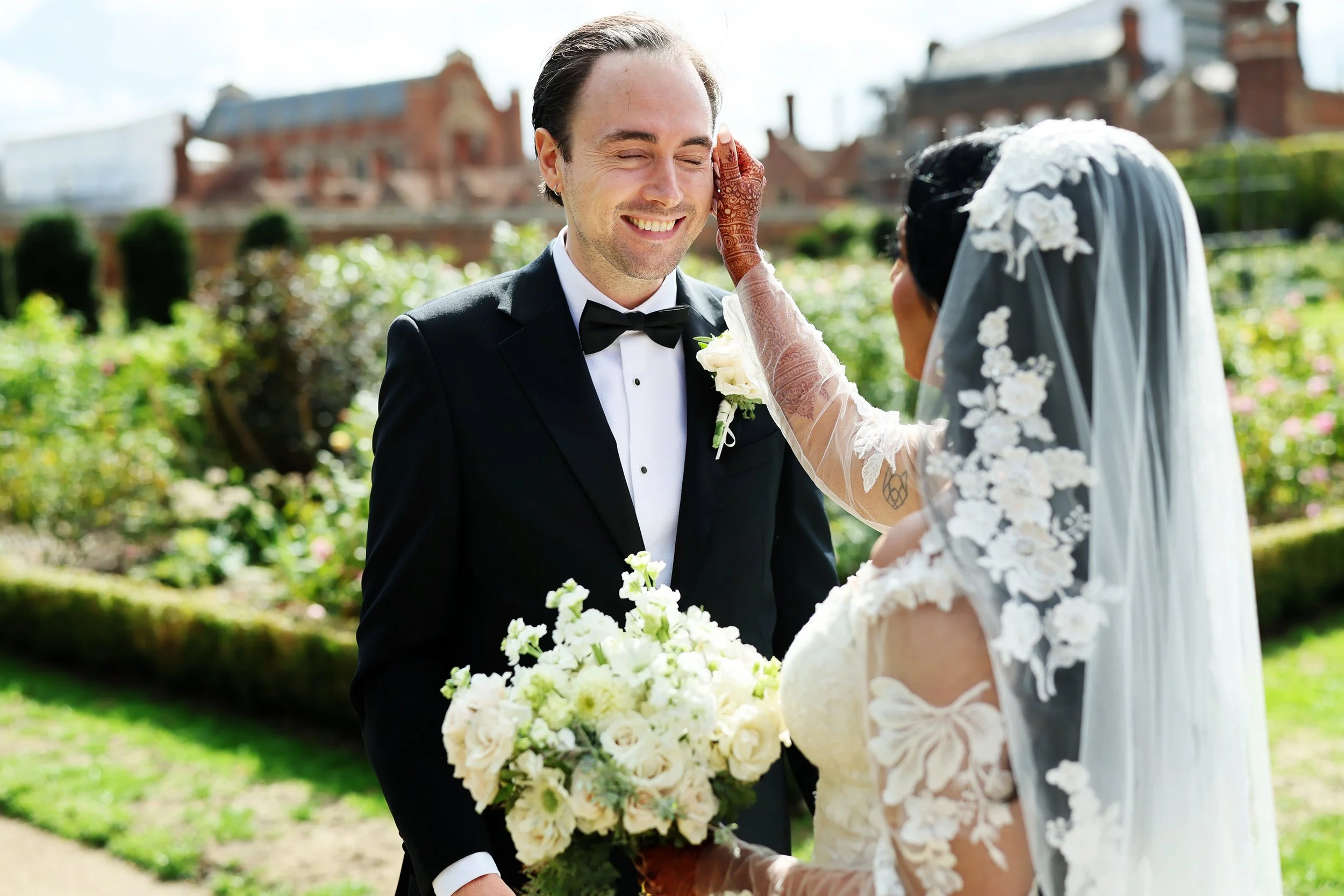 A smiling groom in a black tuxedo with a bow tie stands outside during a wedding ceremony, looking at the bride. The bride is wearing a lace wedding dress and veil, holding a bouquet of white roses and greenery. She is touching the groom's face gentl
