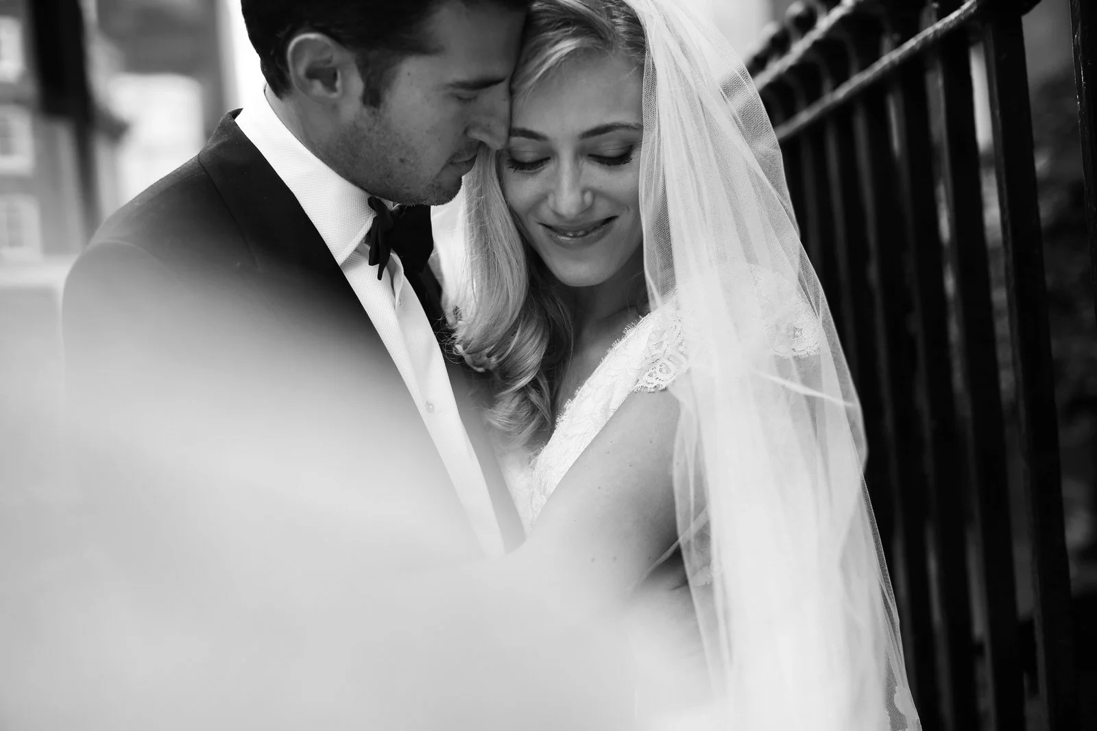 Black and white photo of a bride and groom close together, smiling with their foreheads touching, outdoors near a black fence.