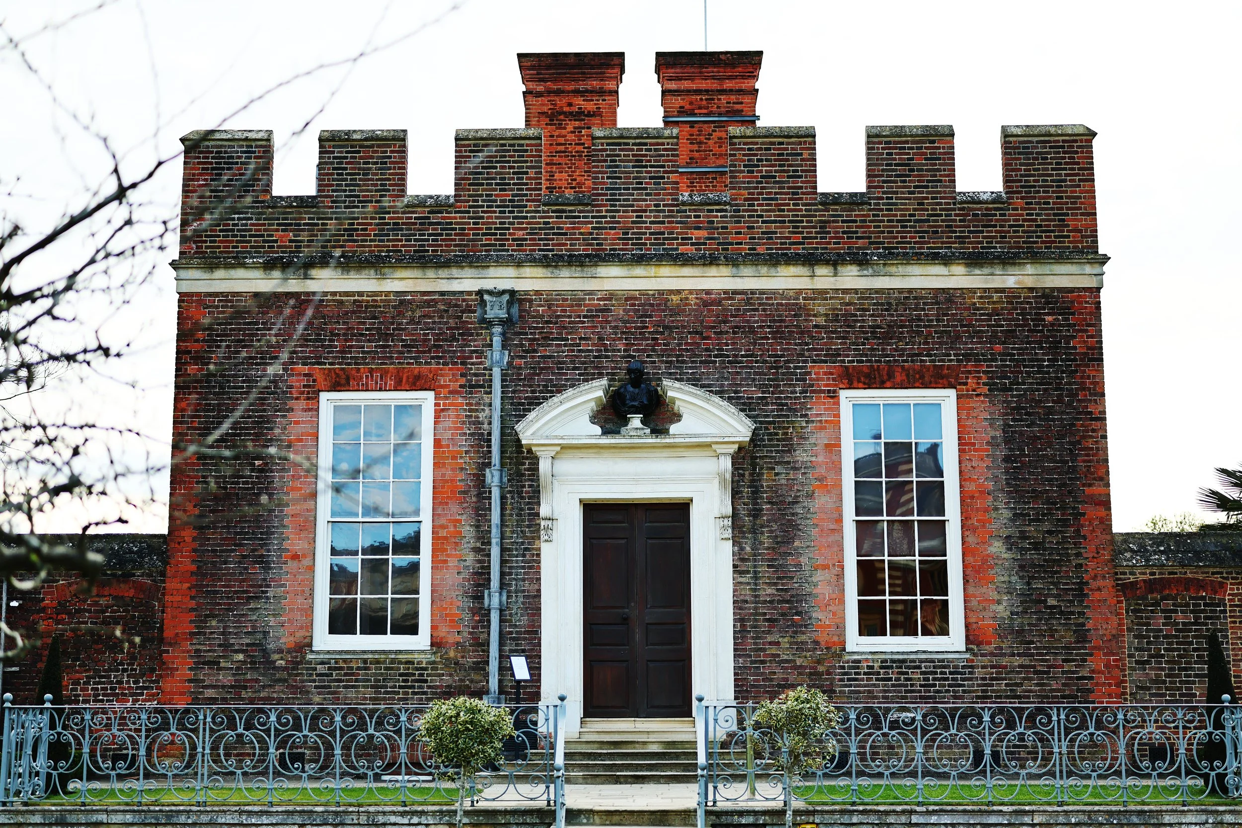 A historic brick building with a black door, large windows, a statue and decorative white trim, surrounded by a blue decorative fence and small plants.
