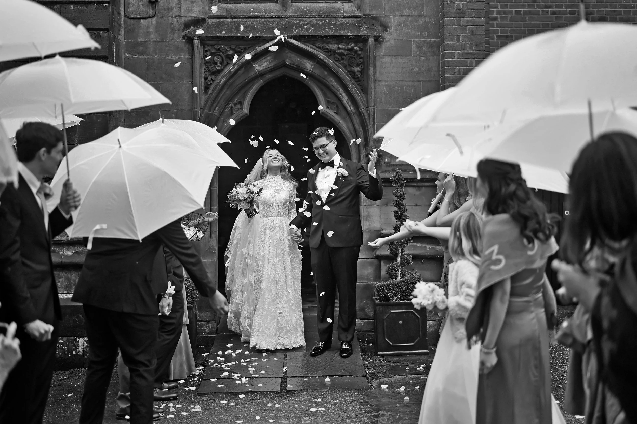 A black and white photo of a newlywed couple walking out of a church, holding hands and smiling as flower petals fall around them. Guests are on both sides under umbrellas.