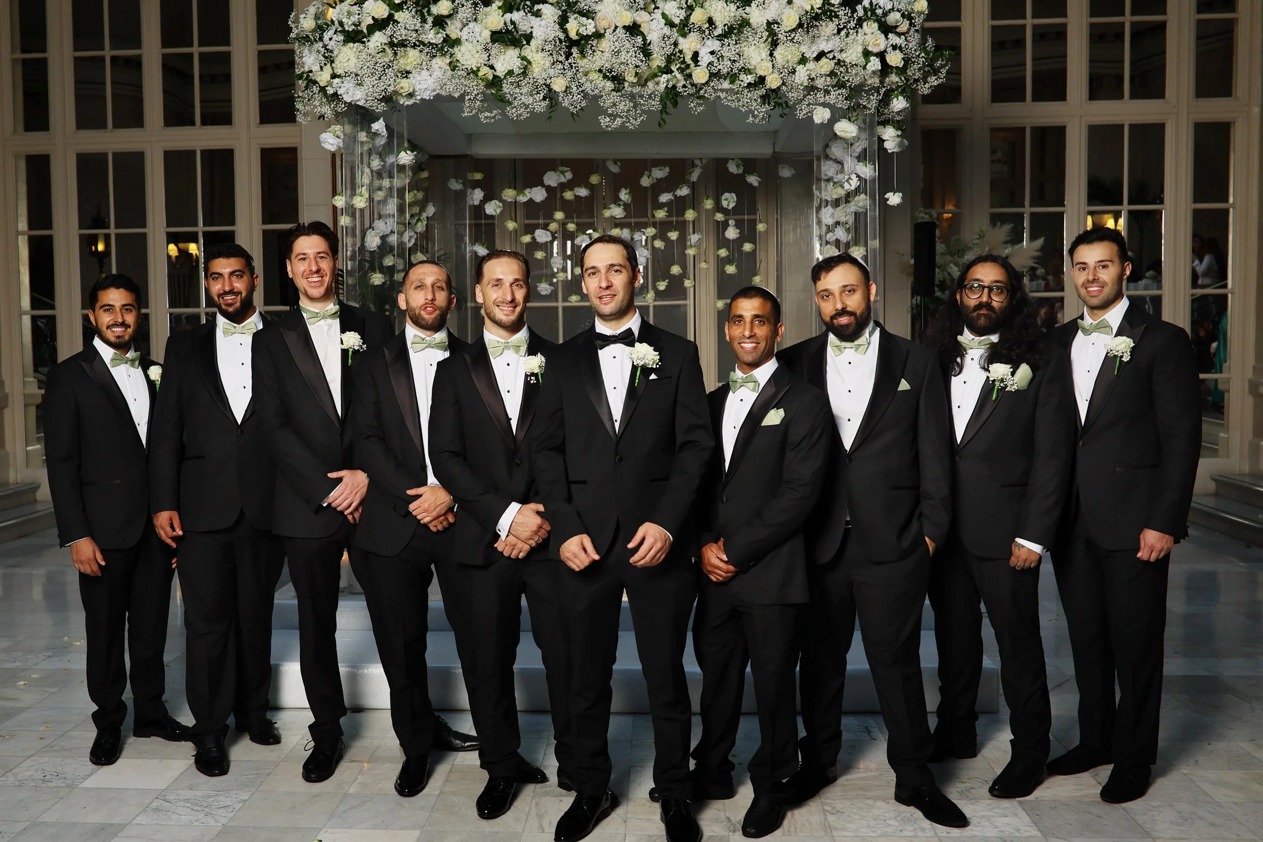Group of eleven men dressed in black tuxedos with light green bow ties, standing in a line indoors with large windows and floral decorations above them.