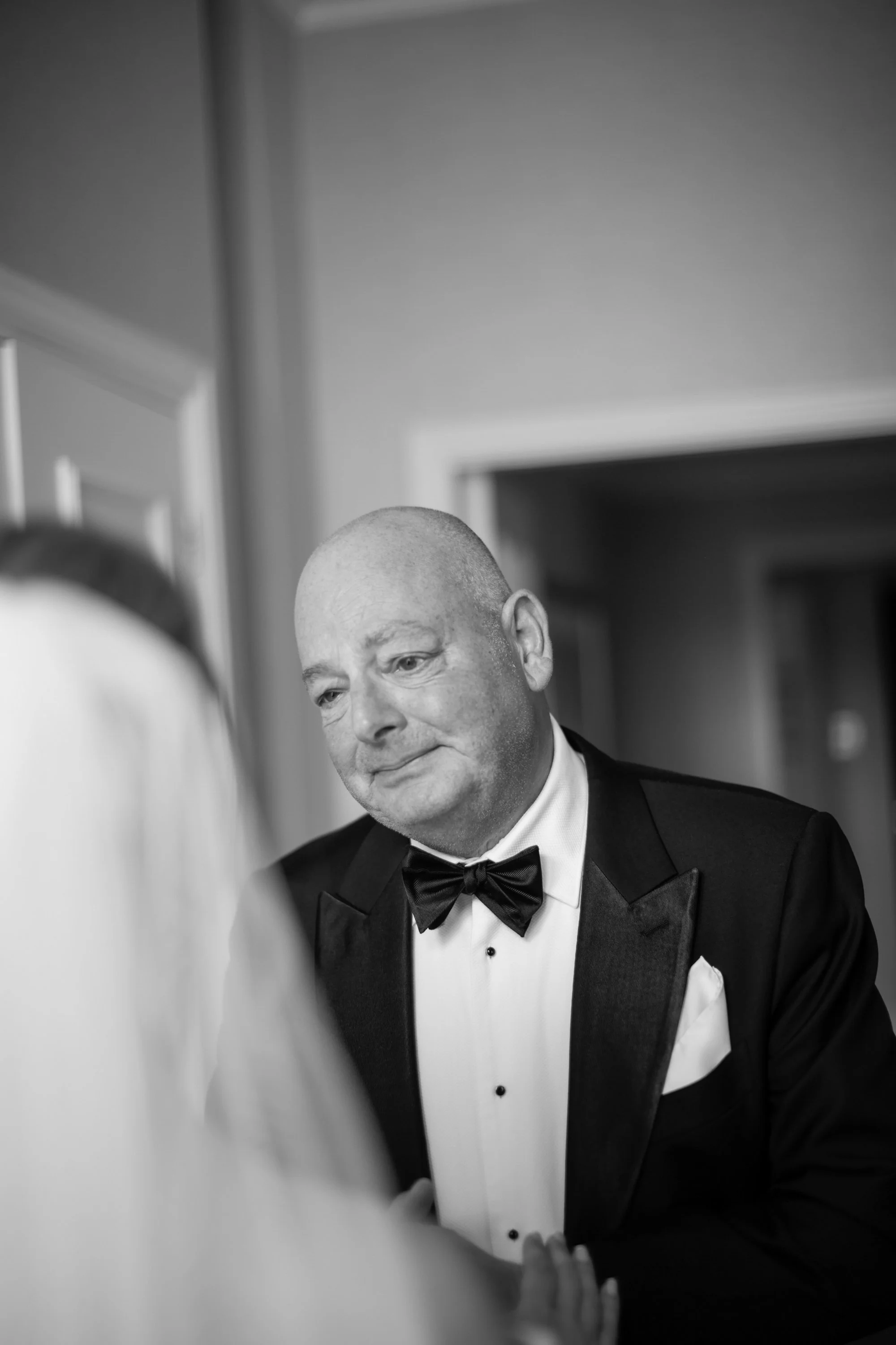 A black-and-white photo of a man in a tuxedo with a bow tie, looking at a mirror, with part of his face and arm visible, and a blurred background.