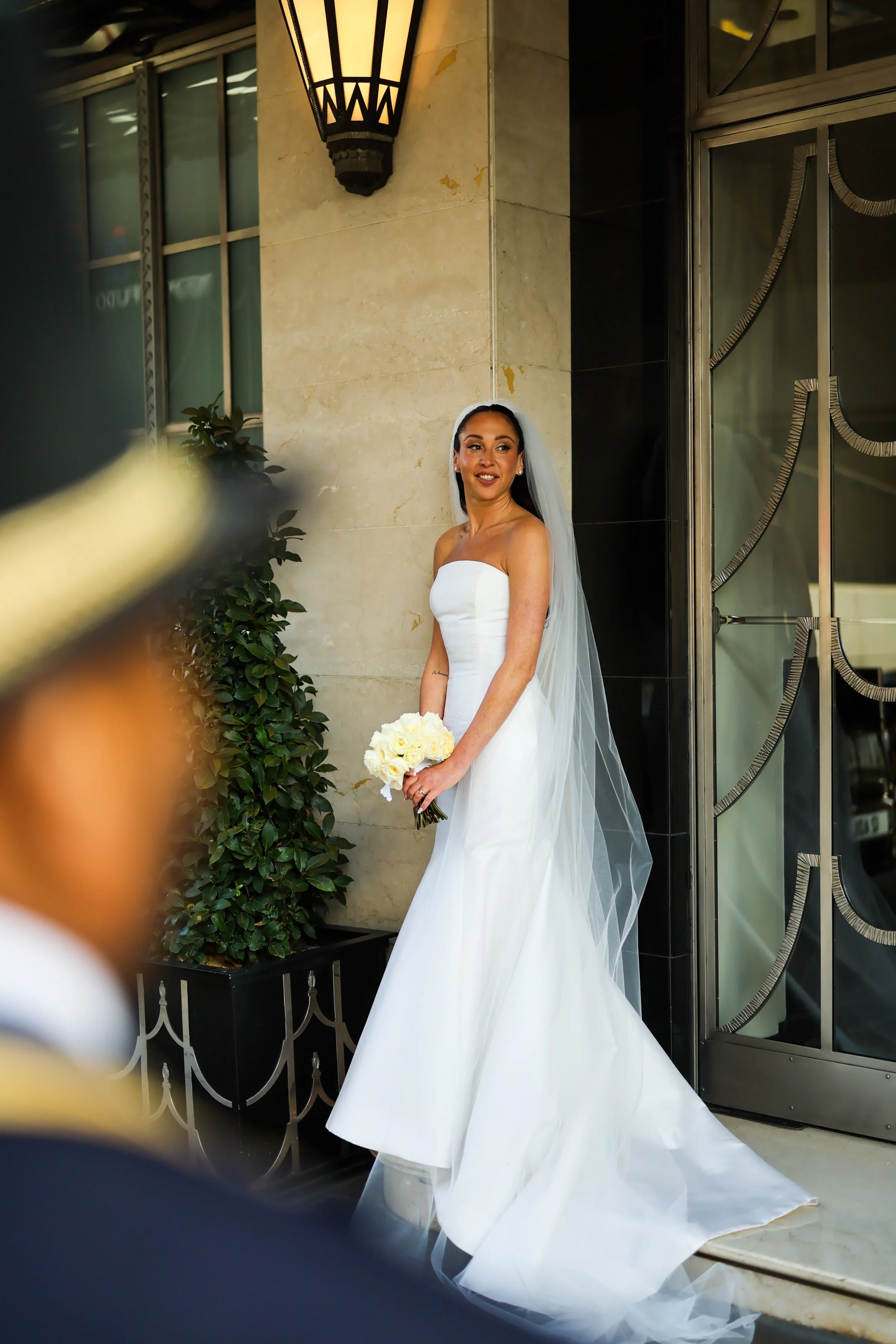 A bride in a strapless white wedding gown with a long veil holding a bouquet of white flowers, standing outside near a beige marble wall and decorative iron gate, with a woman in a floral blouse partially visible in the left foreground.
