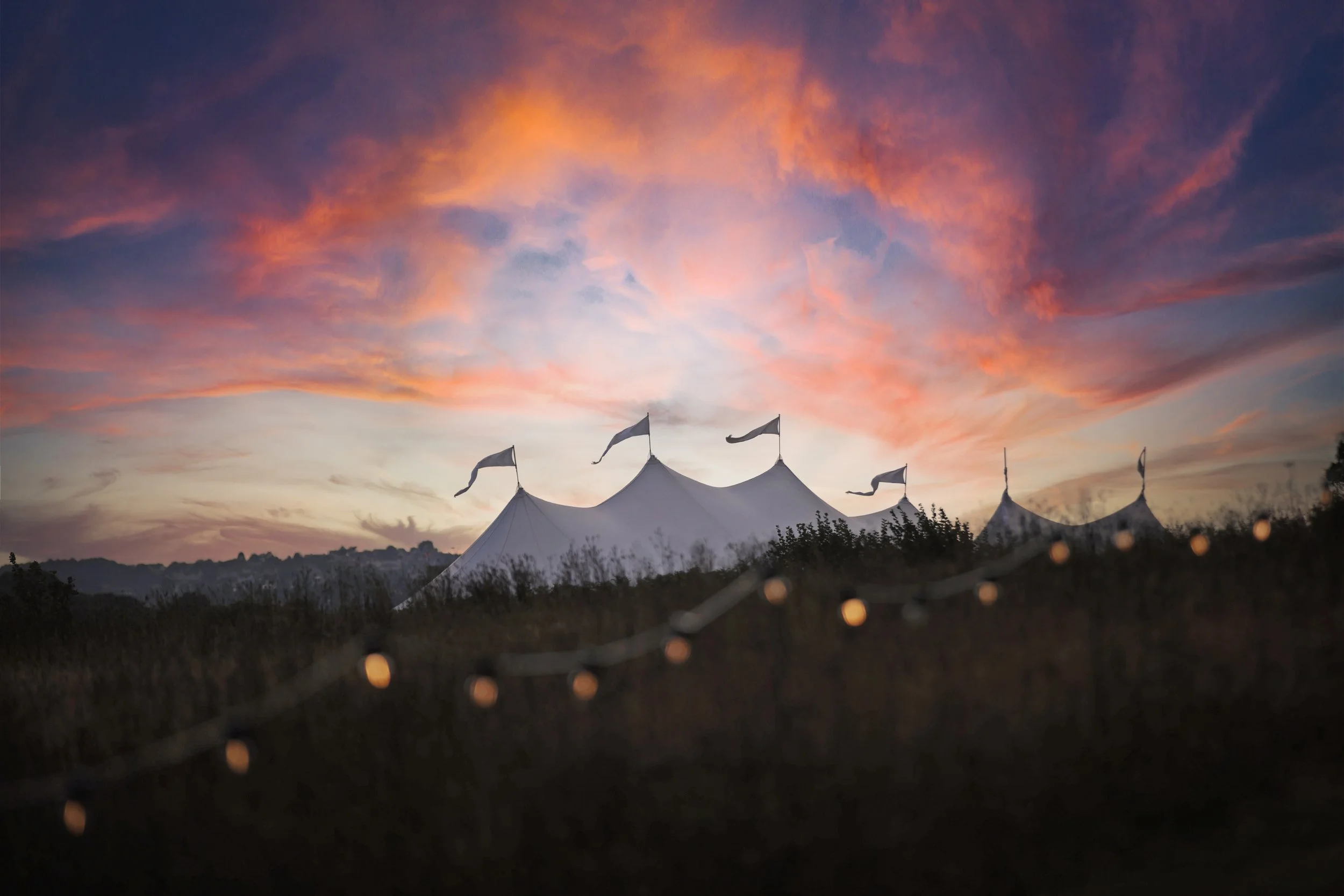 Pink and purple clouds at sunset above white circus tents with flags.