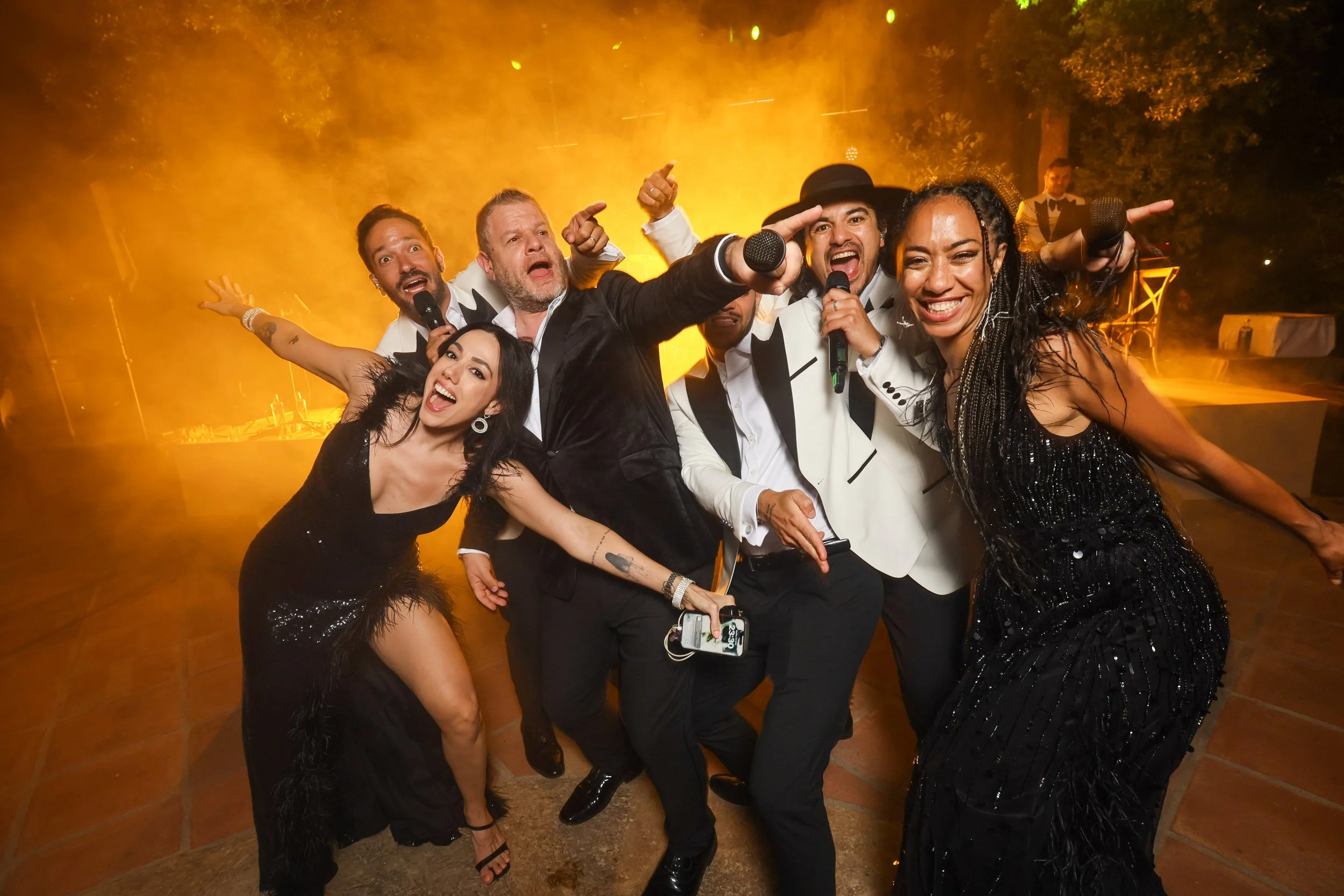 A group of five diverse people dressed in formal and stylish clothes, holding microphones, and singing or partying on a dance floor with orange lighting and fog effects. They appear to be celebrating or performing at a lively event.