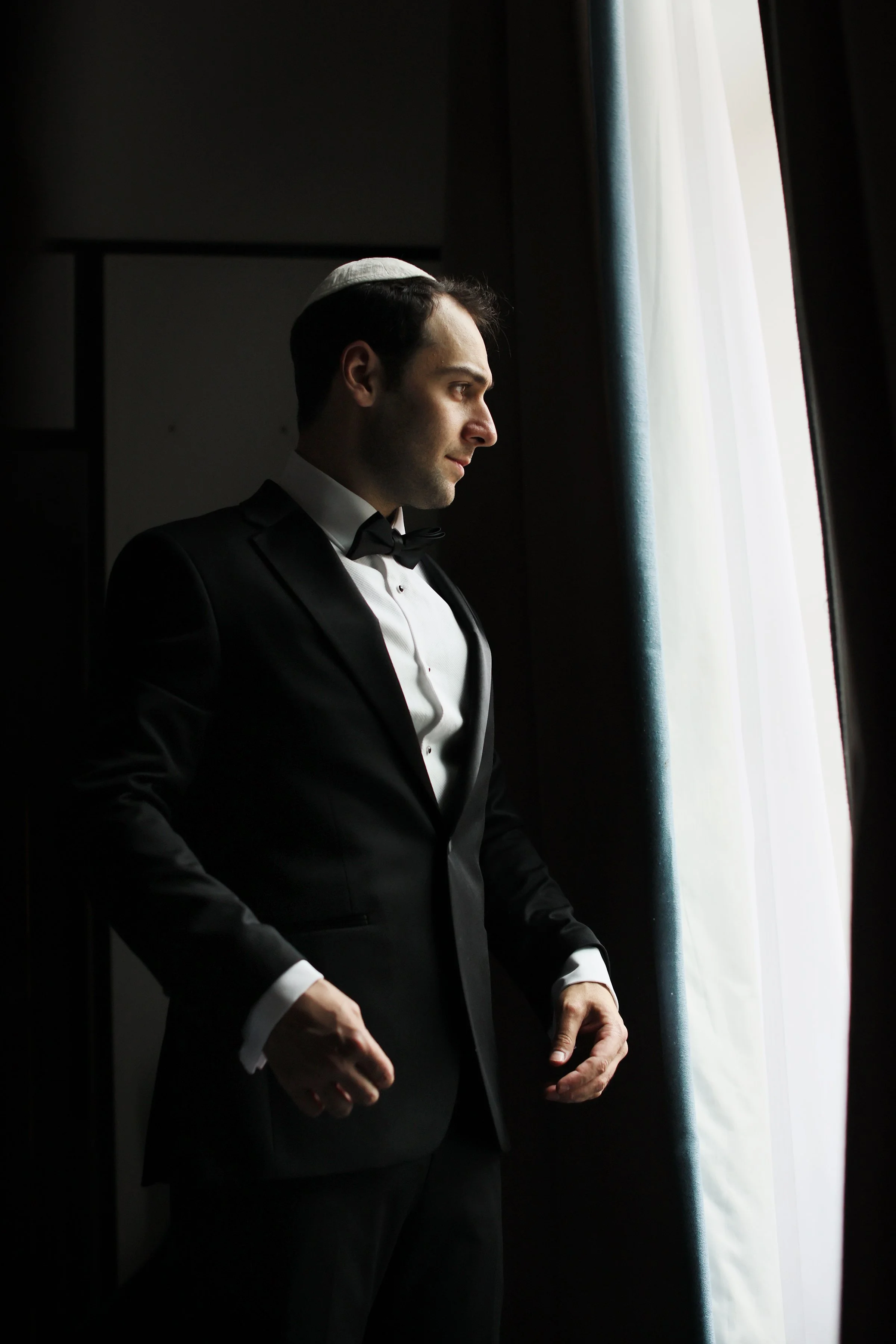 A man in a black tuxedo and bow tie looks out a window with white curtains, wearing a kippah.