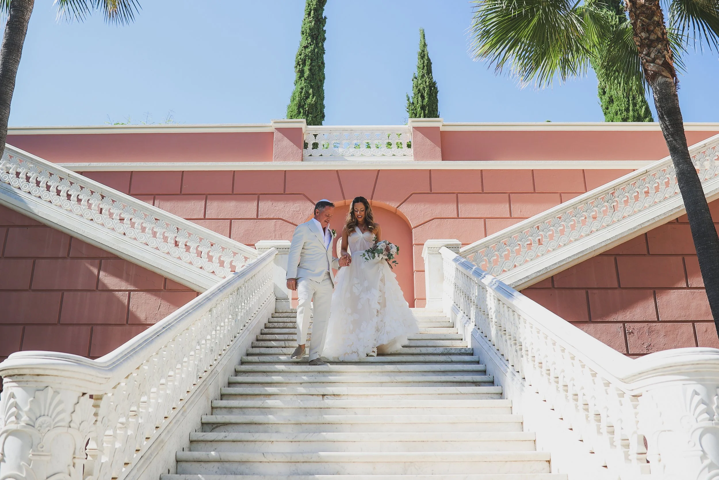 A bride and groom walking down grand outdoor white marble stairs with ornate white railings, against a pink building with classical architectural details, lush greenery, and clear blue sky.