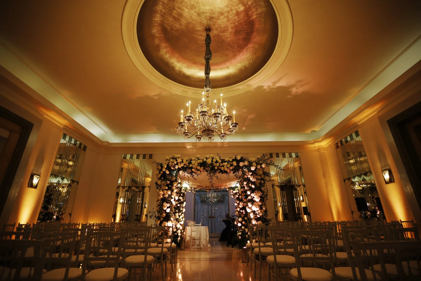 Elegant event space decorated with a floral arch and a chandelier, prepared for a wedding or celebration.