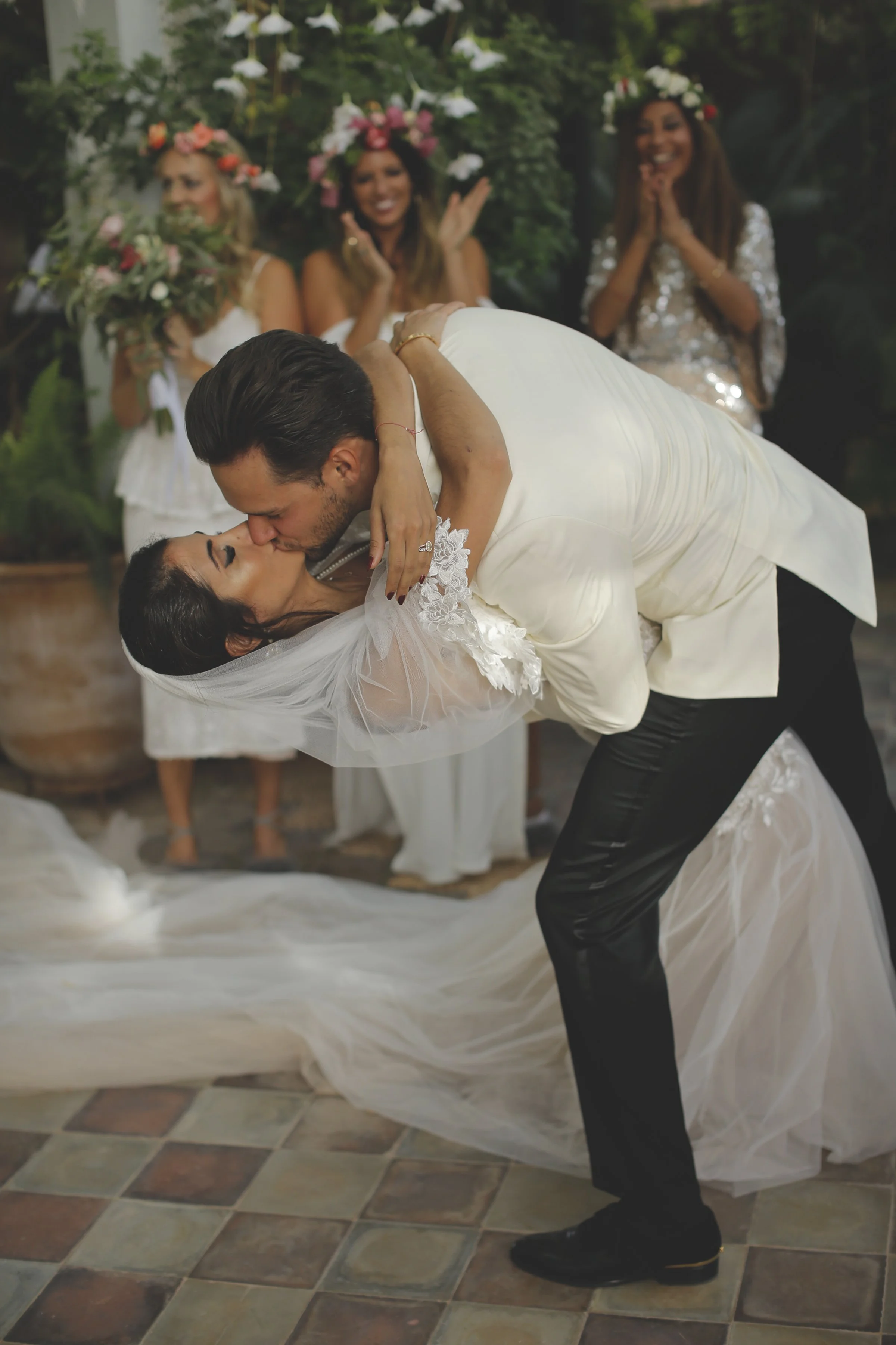 A man in formal attire dips a woman in a wedding dress for a kiss during a wedding celebration. Three women in floral crowns and dresses stand in the background, smiling and clapping, holding bouquets. The setting appears to be an outdoor or garden v