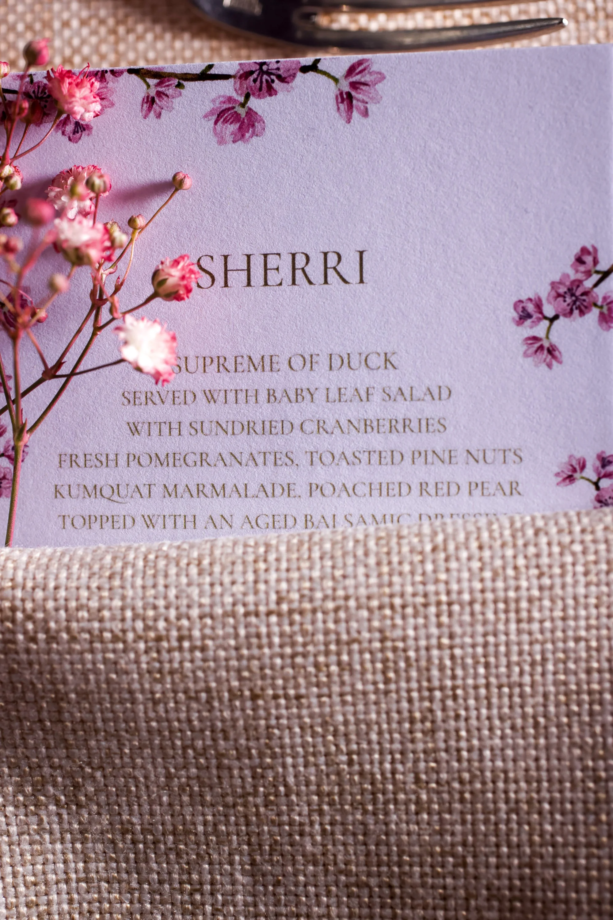 Close-up of a lavender-colored menu card with gold lettering, decorated with pink flowers and placed on a textured pink fabric surface.