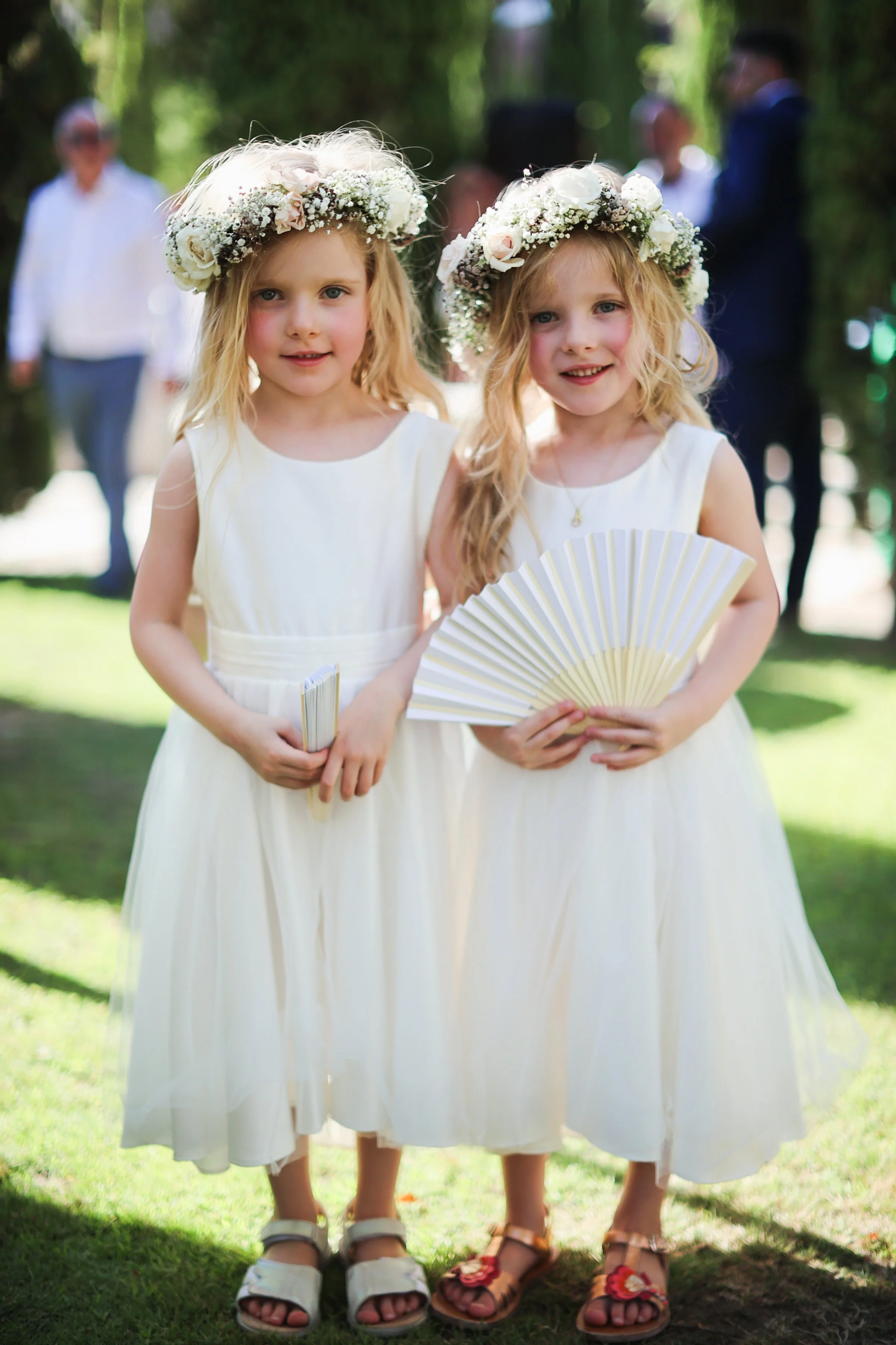 Two young girls wearing white dresses and floral crowns made of white and blush roses stand outdoors in a garden, holding handheld fans. In the background, three adults are visible, slightly blurred, in a shaded area with trees.