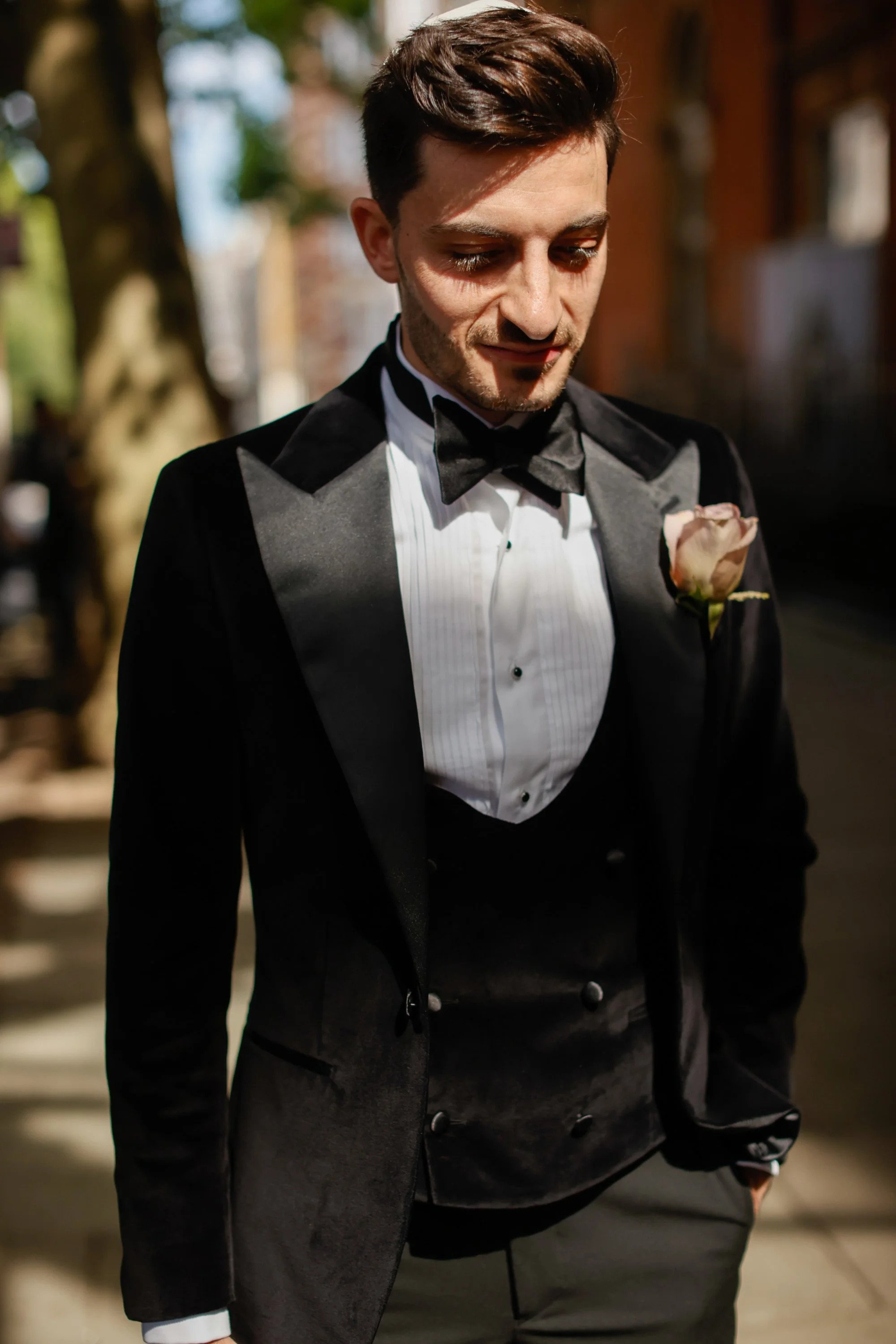 A man dressed in a tuxedo with a white shirt, black bow tie, and a gray waistcoat with a pink rose boutonniere, looking down outdoors with trees and buildings in the background.